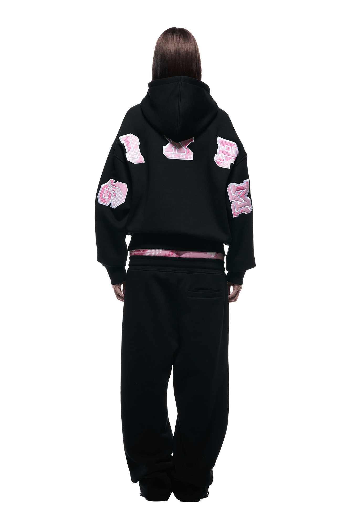 A person with straight brown hair stands with their back to the camera, wearing the 6PM DOT CAMO LETTER HOODIE BLACK. The hoodie features large pink and white varsity-style patches on the sleeves and back.