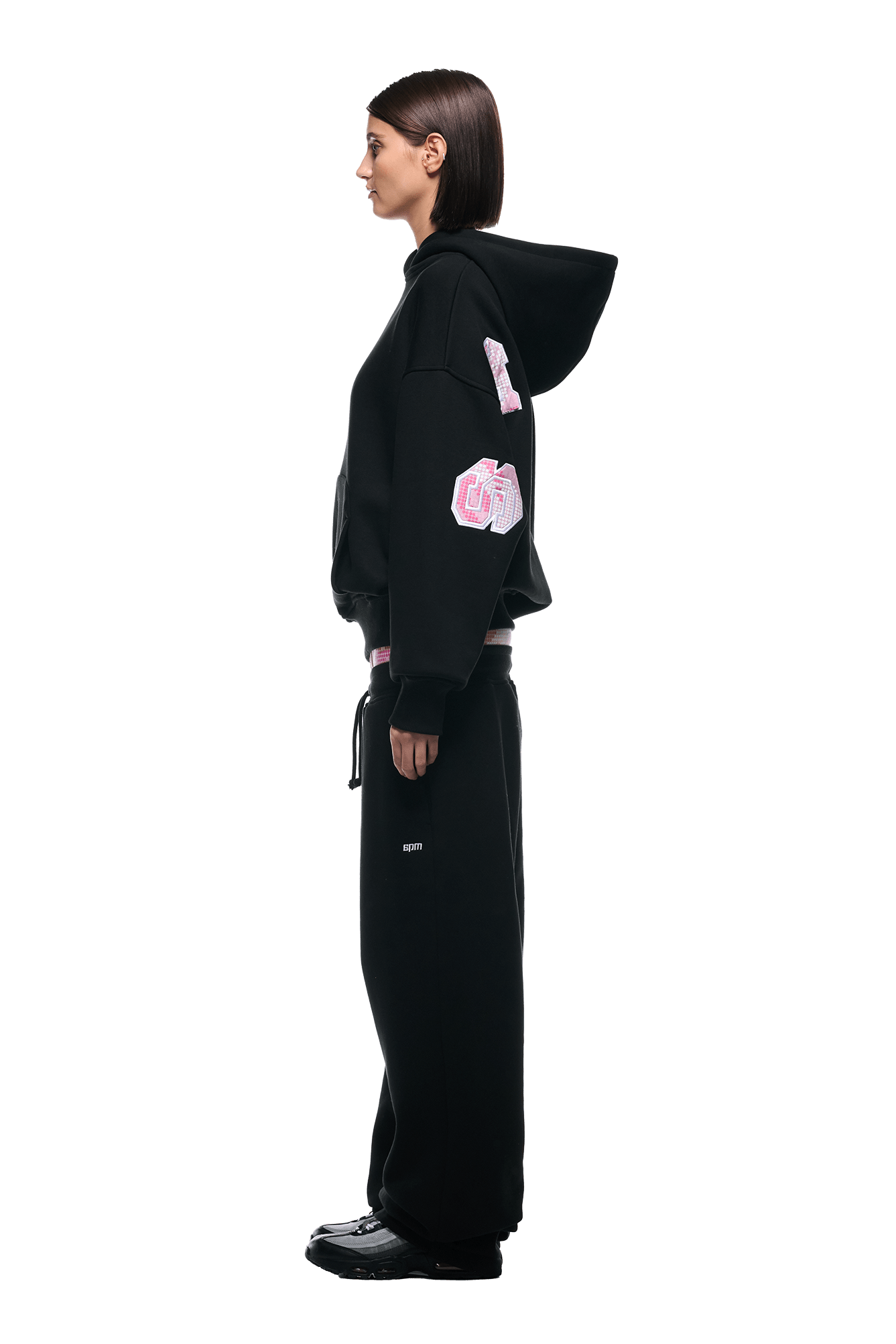 A woman with straight, dark hair wears the 6PM DOT CAMO LETTER HOODIE BLACK and matching black sweatpants, standing in profile against a white background. The hoodie features pink and white patches on the sleeves; she wears black shoes.