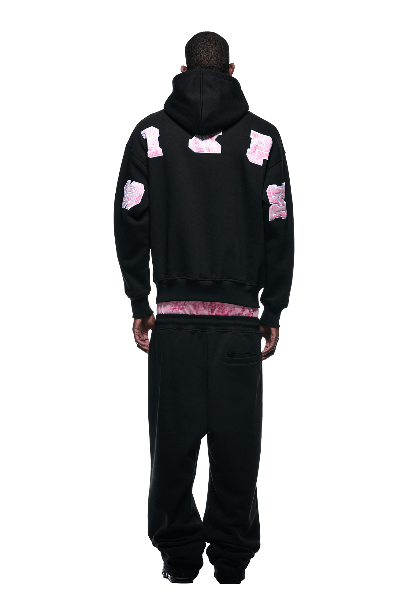 A person stands facing away in a 6PM DOT CAMO LETTER HOODIE BLACK with large pink and white graphic patches on the back and sleeves, paired with loose black pants. The hood is up, and the background is plain white.