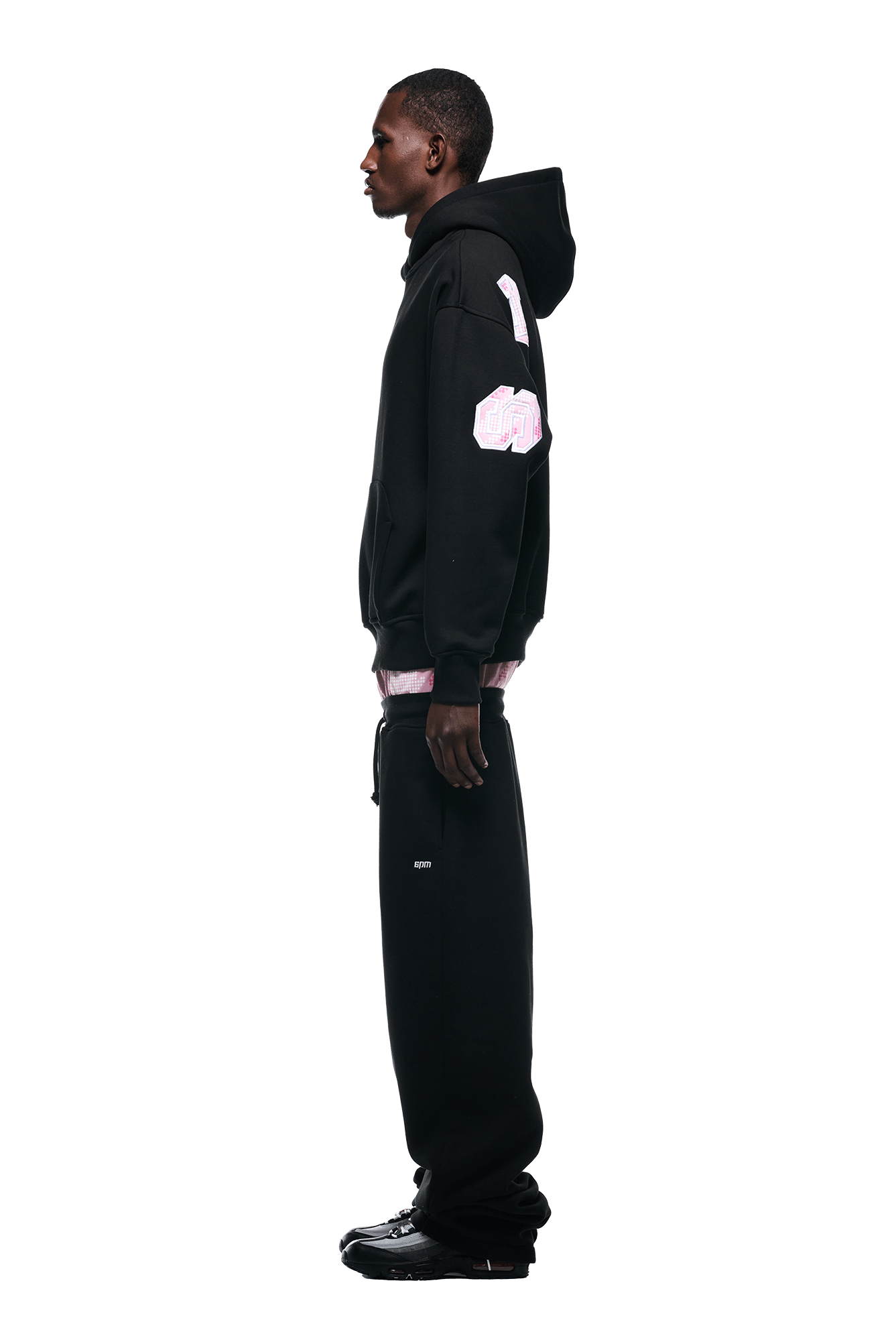 A man stands in profile against a plain white background, wearing the 6PM DOT CAMO LETTER HOODIE BLACK with pink patches, paired with black sweatpants and black sneakers.