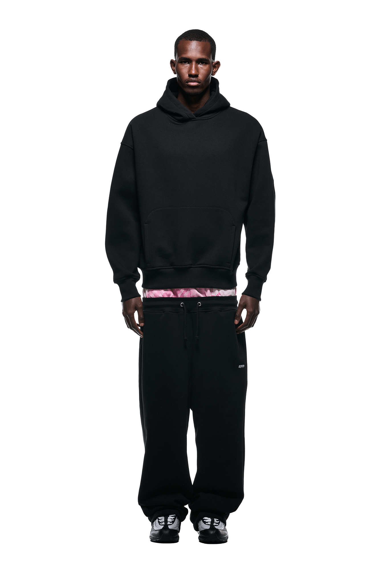 A person stands facing forward in a 6PM DOT CAMO LETTER HOODIE BLACK over a pink tie-dye shirt, paired with black sweatpants and black sneakers. The background is plain white.