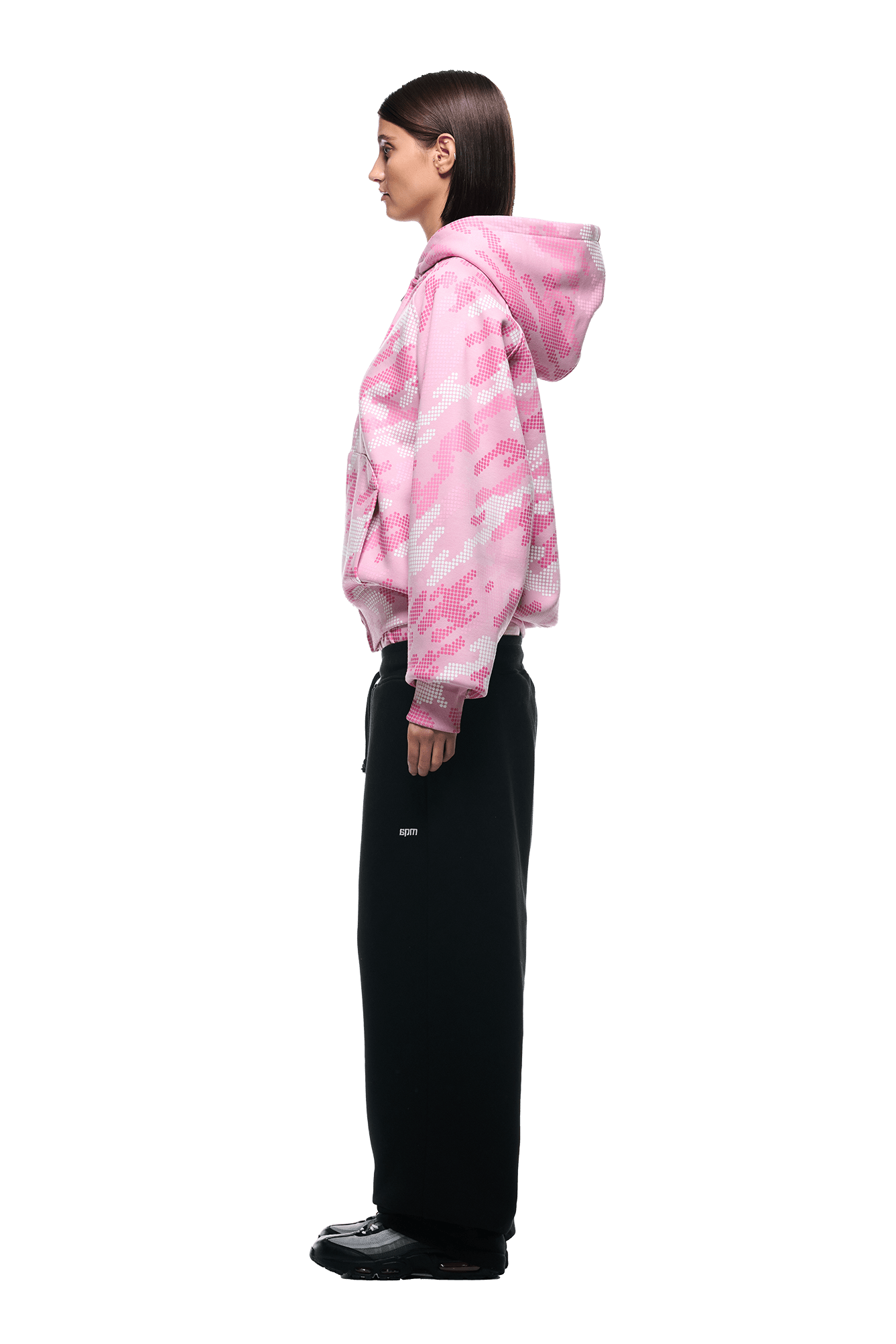 A woman stands in profile against a plain white background, wearing the 6PM DOT CAMO ZIP HOODIE PINK with the hood down, paired with loose black pants and black shoes.