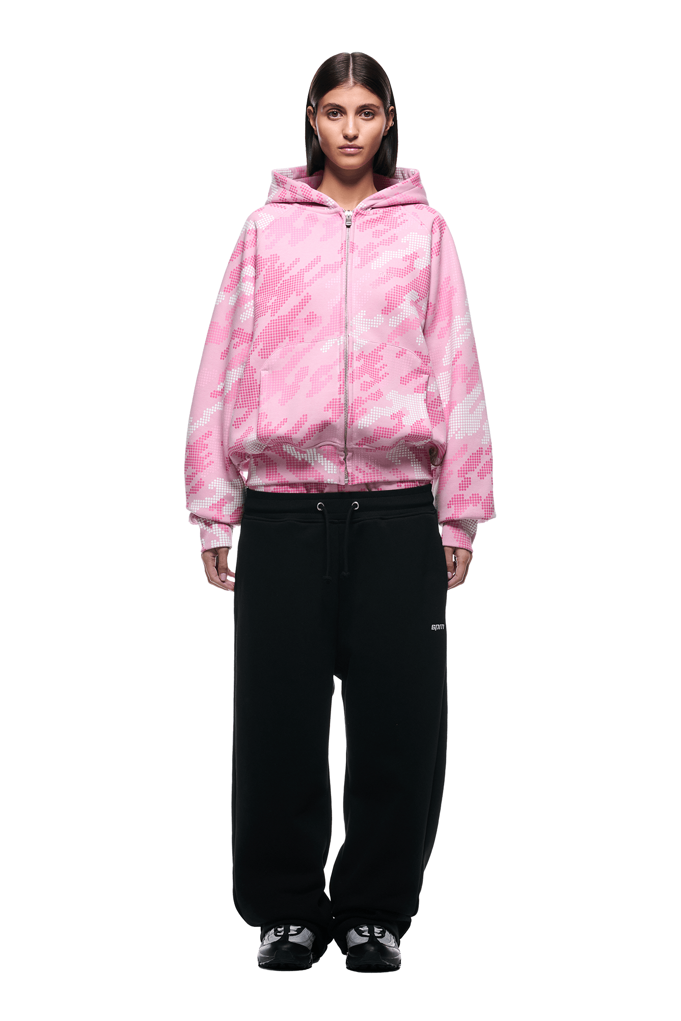 A person stands facing forward against a plain white background, wearing the 6PM DOT CAMO ZIP HOODIE PINK, loose black sweatpants, and black sneakers.