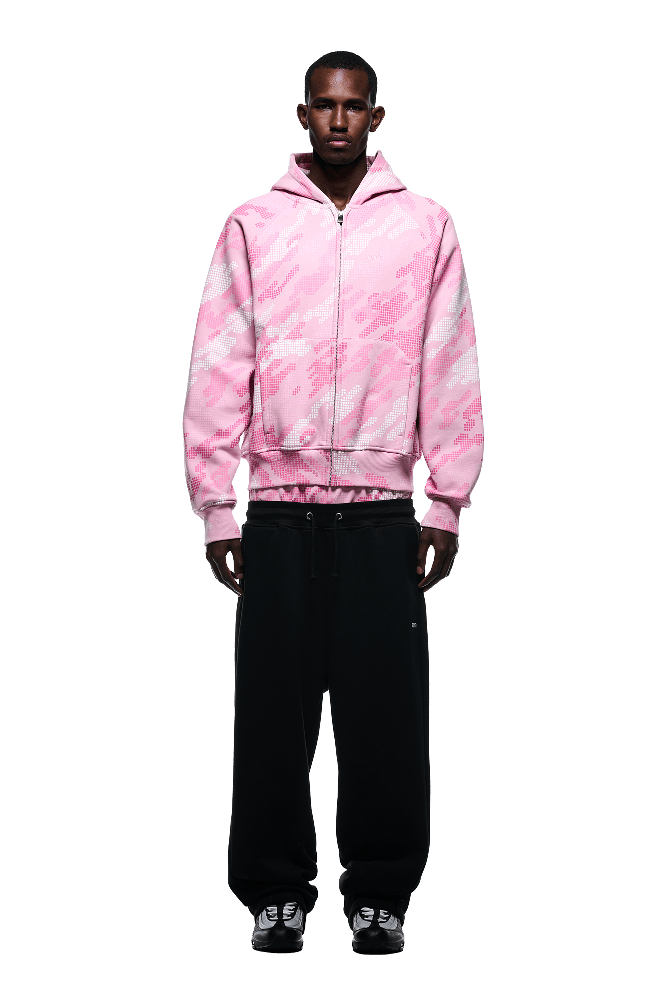 A man faces forward against a white background, wearing the 6PM DOT CAMO ZIP HOODIE PINK, loose black pants, and black sneakers.