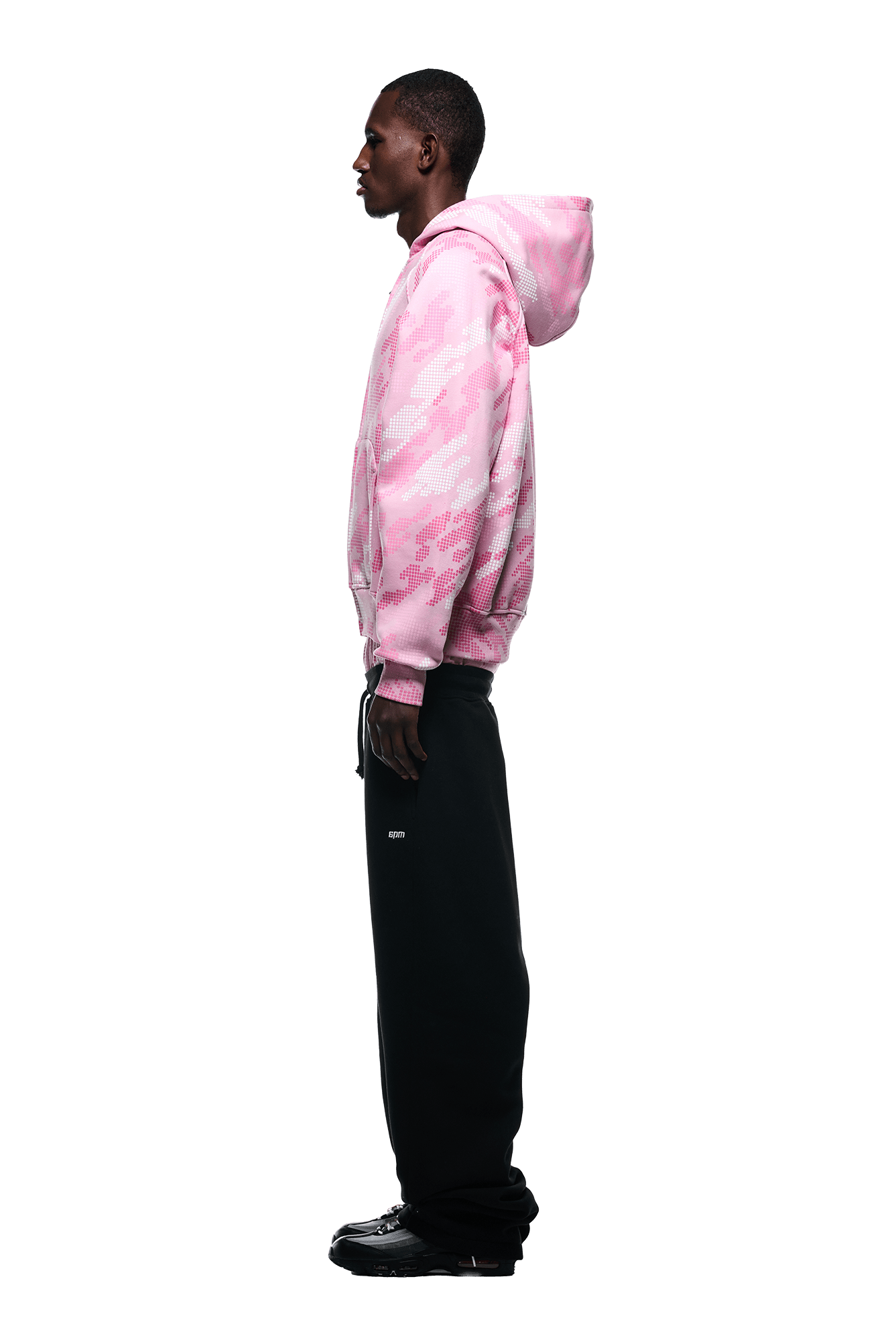 A man stands in profile against a white background, wearing the 6PM DOT CAMO ZIP HOODIE PINK with black wide-leg pants and black shoes.