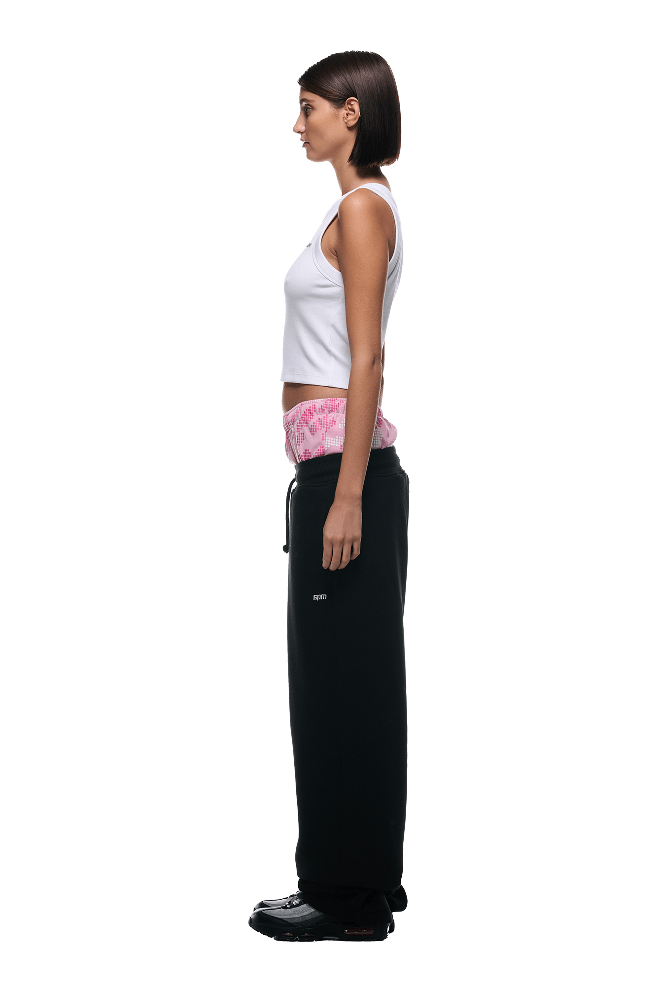 A woman stands in profile against a white background, wearing the 6PM BOXERS OPEN LEG JOGGER BLACK over pink shorts and a white tank top, paired with black shoes. Her straight hair is chin-length.