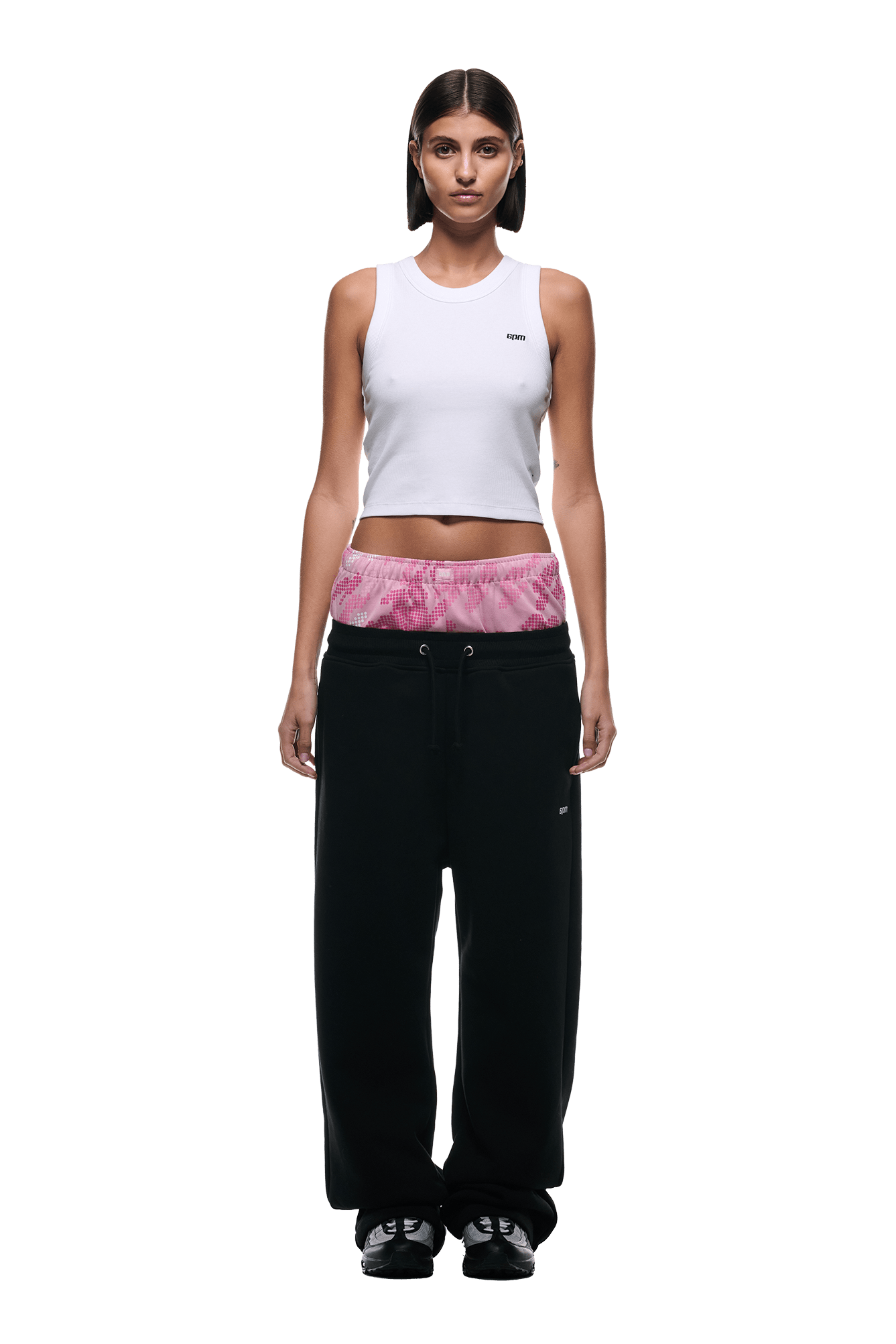A woman with straight dark hair wears a white sleeveless crop top and 6PM BOXERS OPEN LEG JOGGER BLACK pants featuring a pink tie-dye waistband, along with black shoes, standing against a plain white background.