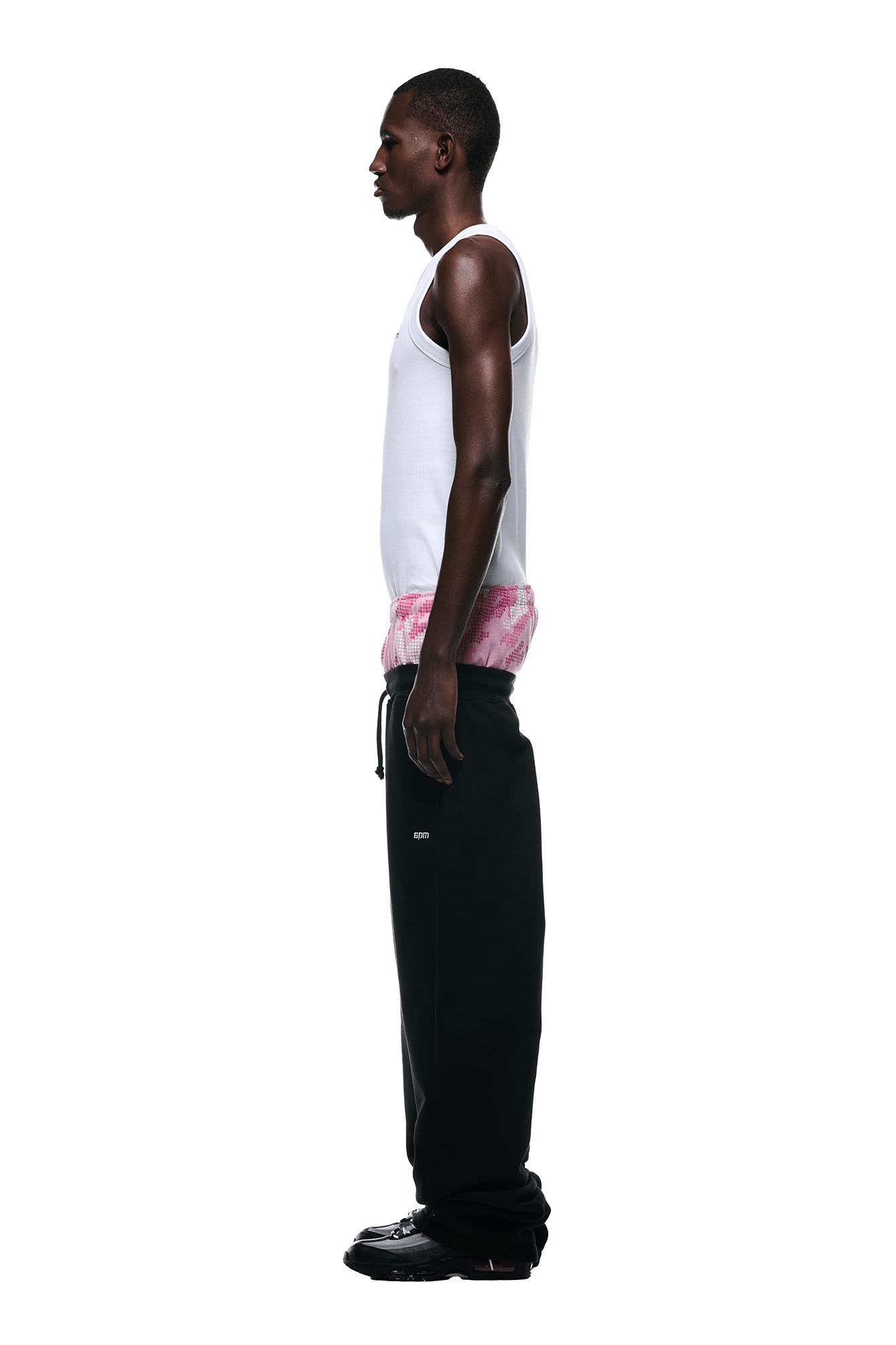A man stands in profile against a white background, sporting 6PMs BOXERS OPEN LEG JOGGER BLACK pants with a drawstring, paired with a white tank top, pink tie-dye underwear, and black shoes.