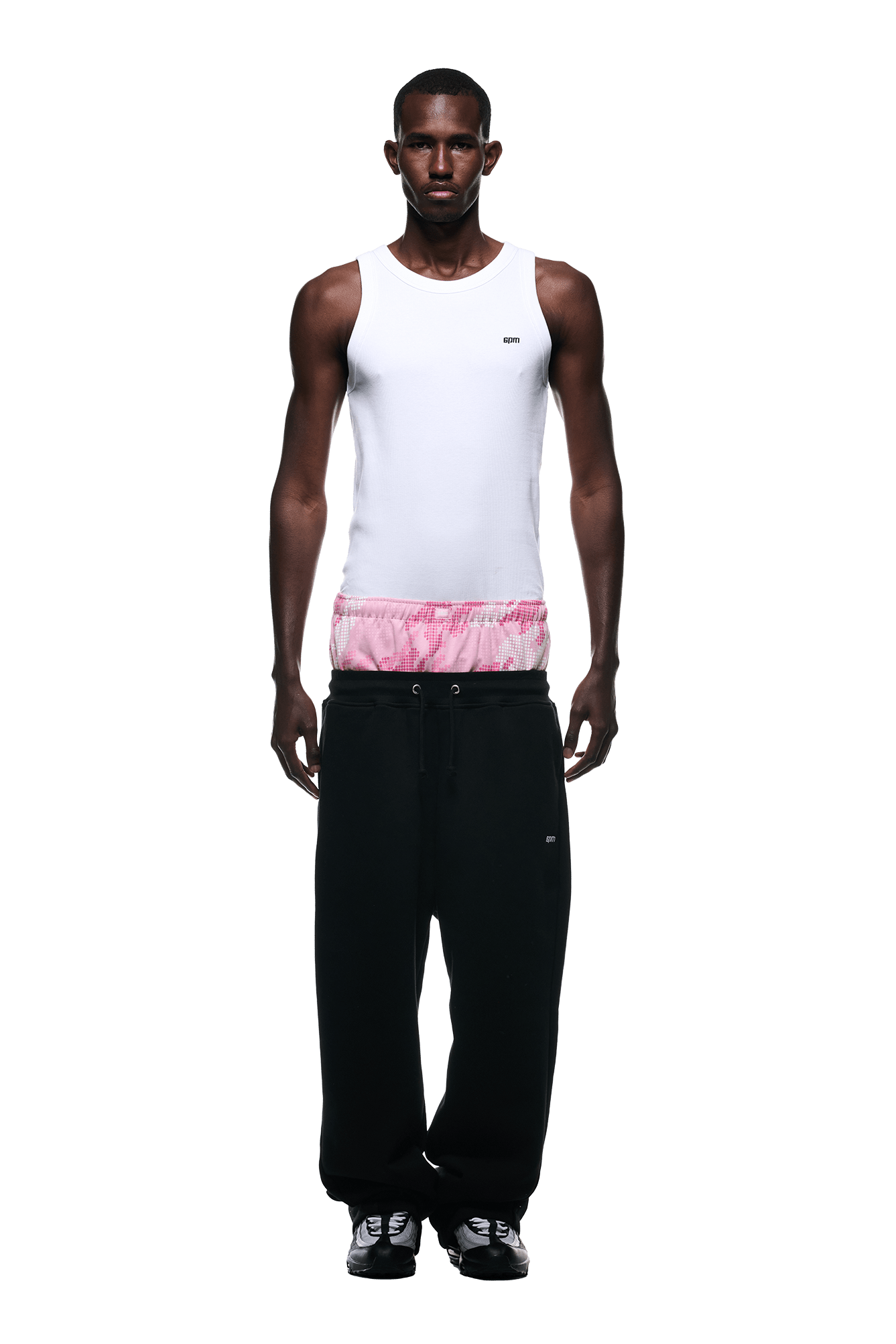 A man stands facing forward against a white background, wearing 6PMs BOXERS OPEN LEG JOGGER BLACK—oversized black pants with a drawstring—paired with a white tank top, pink and white tie-dye shorts, and black sneakers.