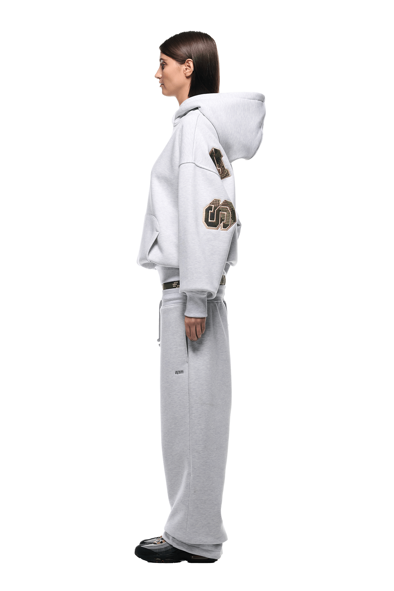 A woman in profile models the 6PM DOT CAMO LETTER HOODIE GREY with matching wide-leg sweatpants. She has straight brown hair, black shoes, and stands against a plain white background.