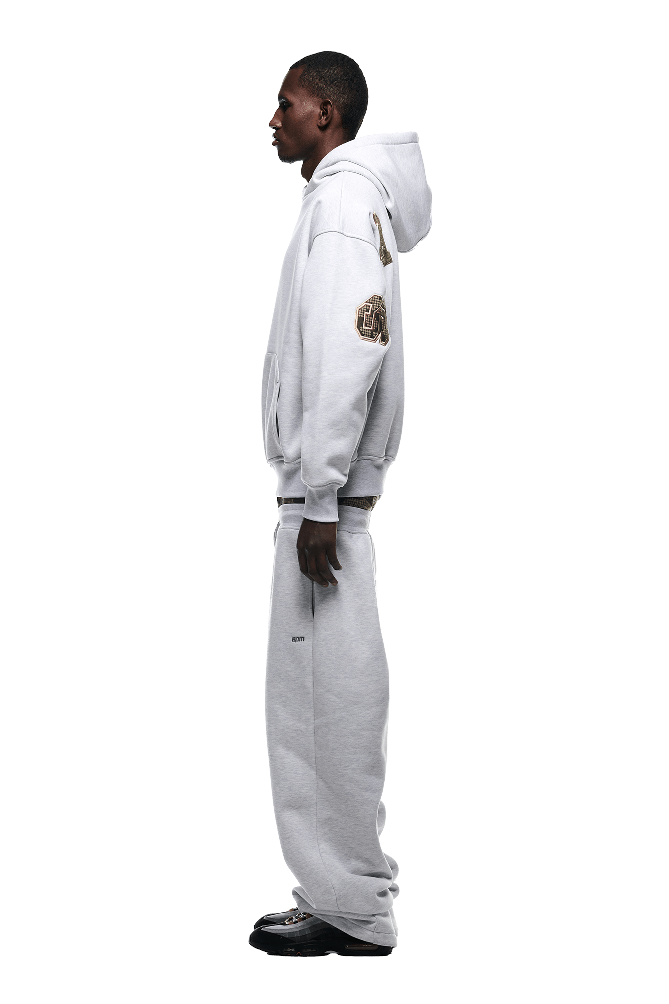 A man with dark skin and short hair stands in profile on a plain white background, wearing the 6PM DOT CAMO LETTER HOODIE GREY with patches on the sleeve, matching loose sweatpants, and black sneakers.