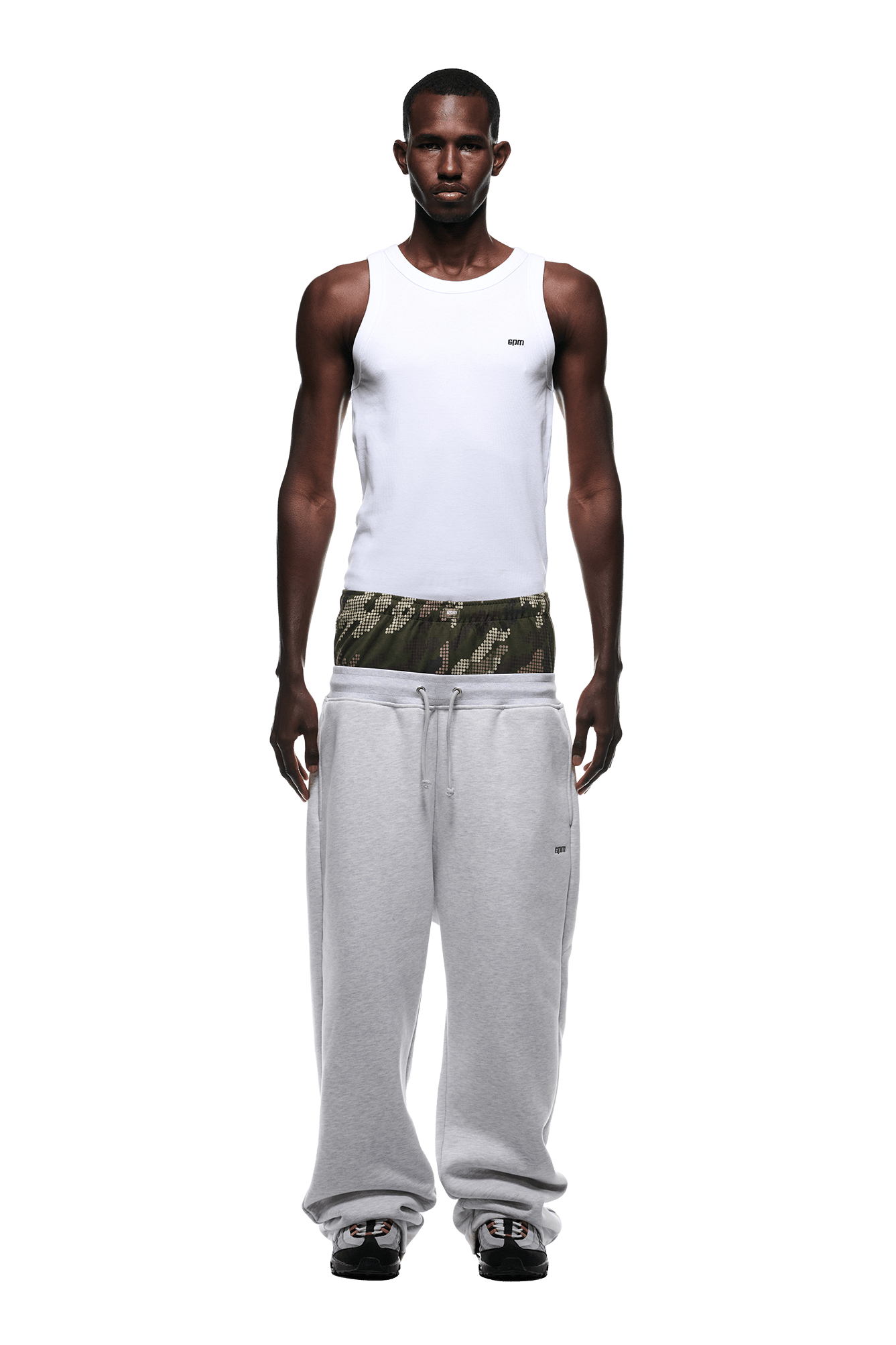 A man faces forward against a plain green background, wearing a white tank top, camouflage boxers, 6PM BOXERS OPEN LEG JOGGER GREY sweatpants with a drawstring, and black-and-white sneakers.