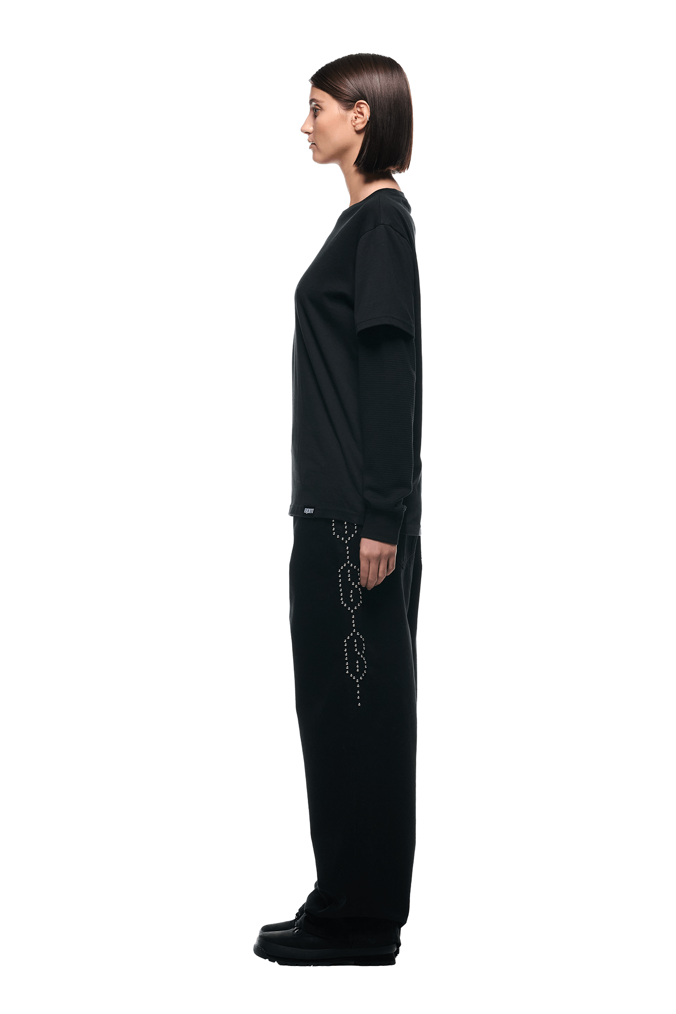 A woman with straight brown hair stands in profile against a white background, wearing the 6PM LAYERED LONGSLEEVE BLACK, black beaded pants, and black shoes.