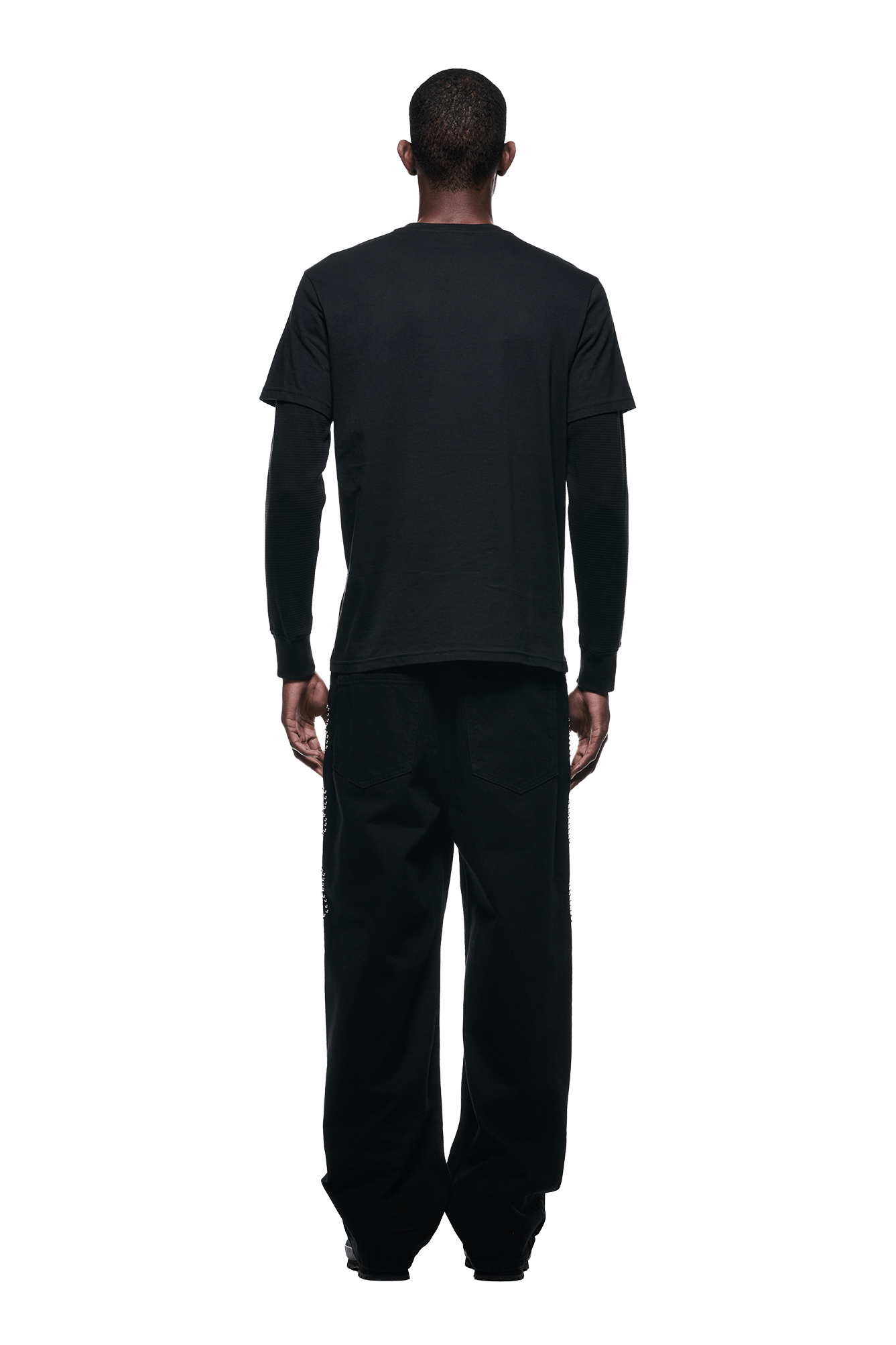 A person faces away from the camera, wearing 6PMs LAYERED LONGSLEEVE BLACK shirt and loose black pants against a plain white background.