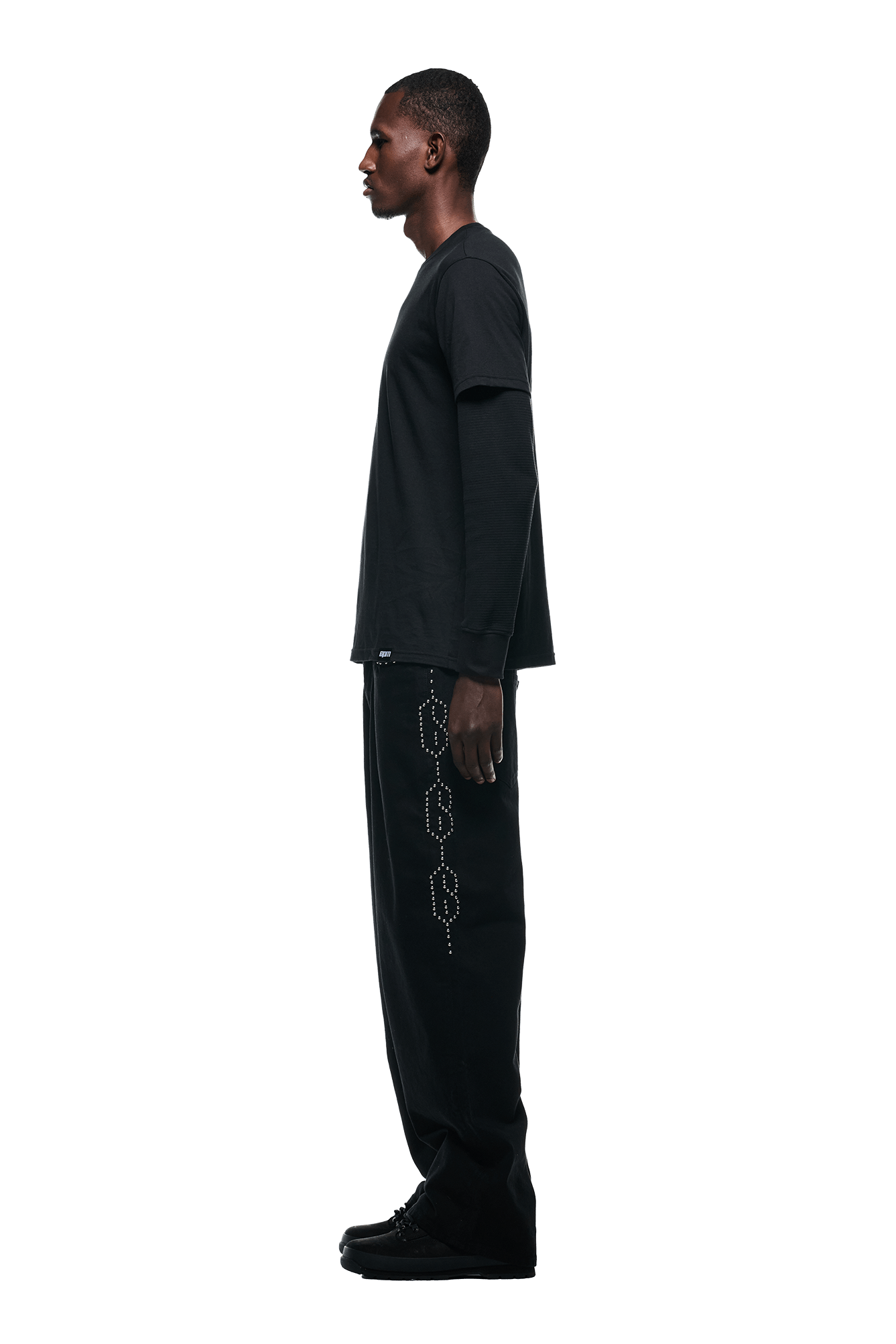A man stands in profile against a white background, wearing the 6PM LAYERED LONGSLEEVE BLACK shirt, black pants with a circular side pattern, and black shoes.