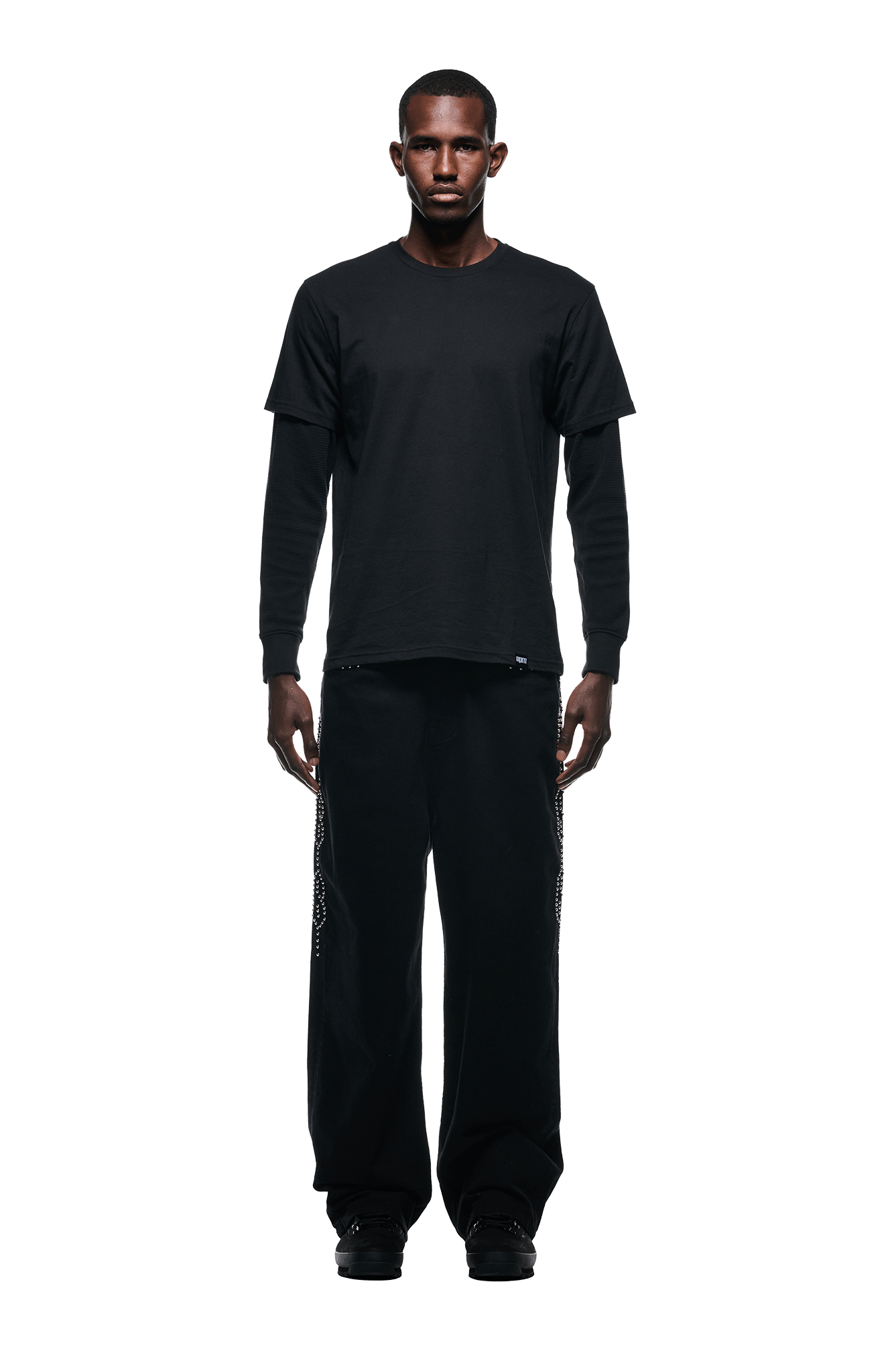 A man faces forward against a white background, wearing the 6PM LAYERED LONGSLEEVE BLACK shirt, loose black pants with side detailing, and black shoes.