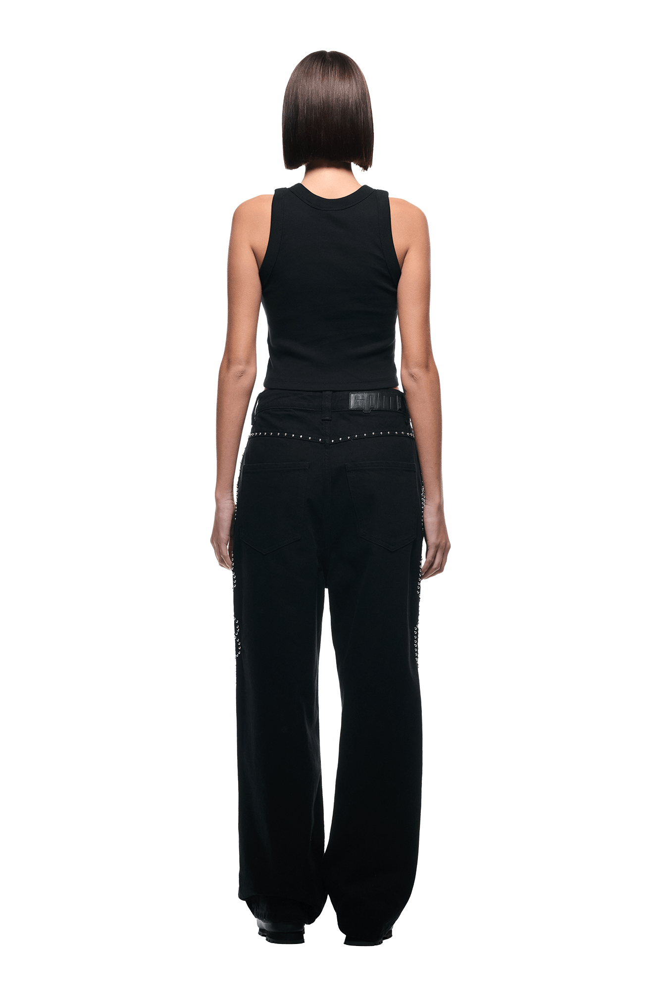 A person with straight, chin-length brown hair faces away, wearing 6PM’s black sleeveless top and STUDS DENIM BLACK pants with studded details, set against a plain white background.