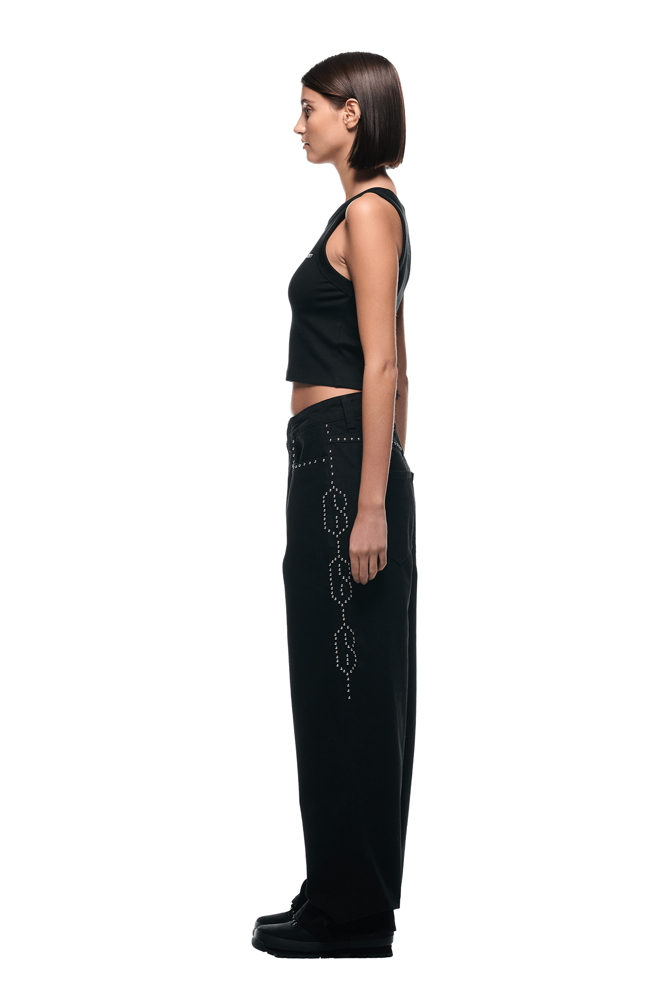 A woman with straight dark hair stands in profile against a white background, wearing 6PM’s STUDS DENIM BLACK high-waisted, wide-leg pants with rhinestone embellishments, a black sleeveless crop top, and black shoes.