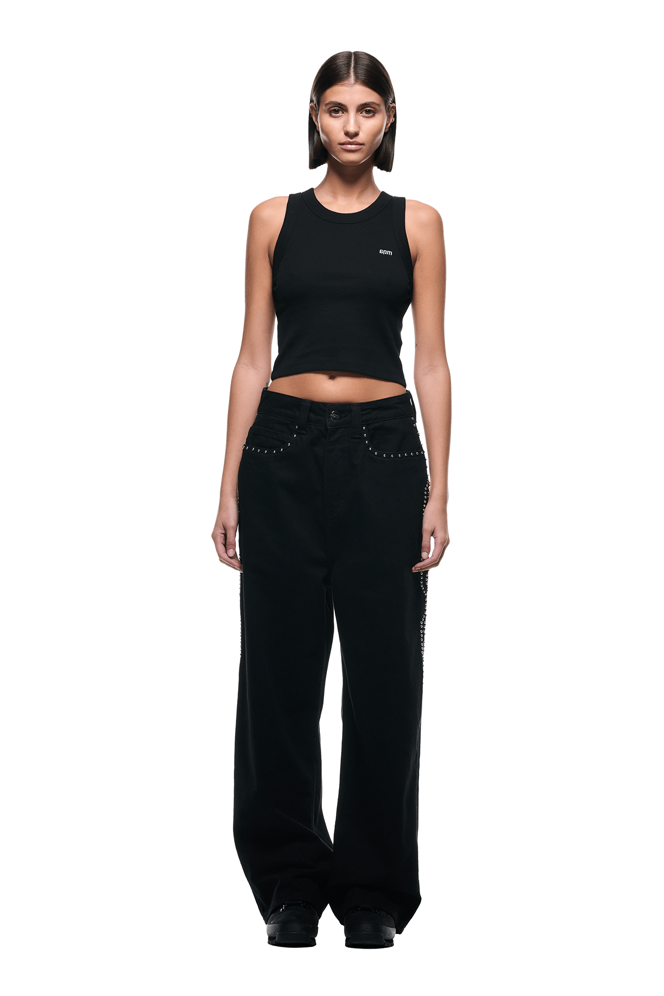 A woman faces forward wearing 6PMs STUDS DENIM BLACK—black, loose-fit pants with stud details—paired with a black sleeveless crop top and black shoes. Her straight, shoulder-length brown hair is styled simply against a plain white background.