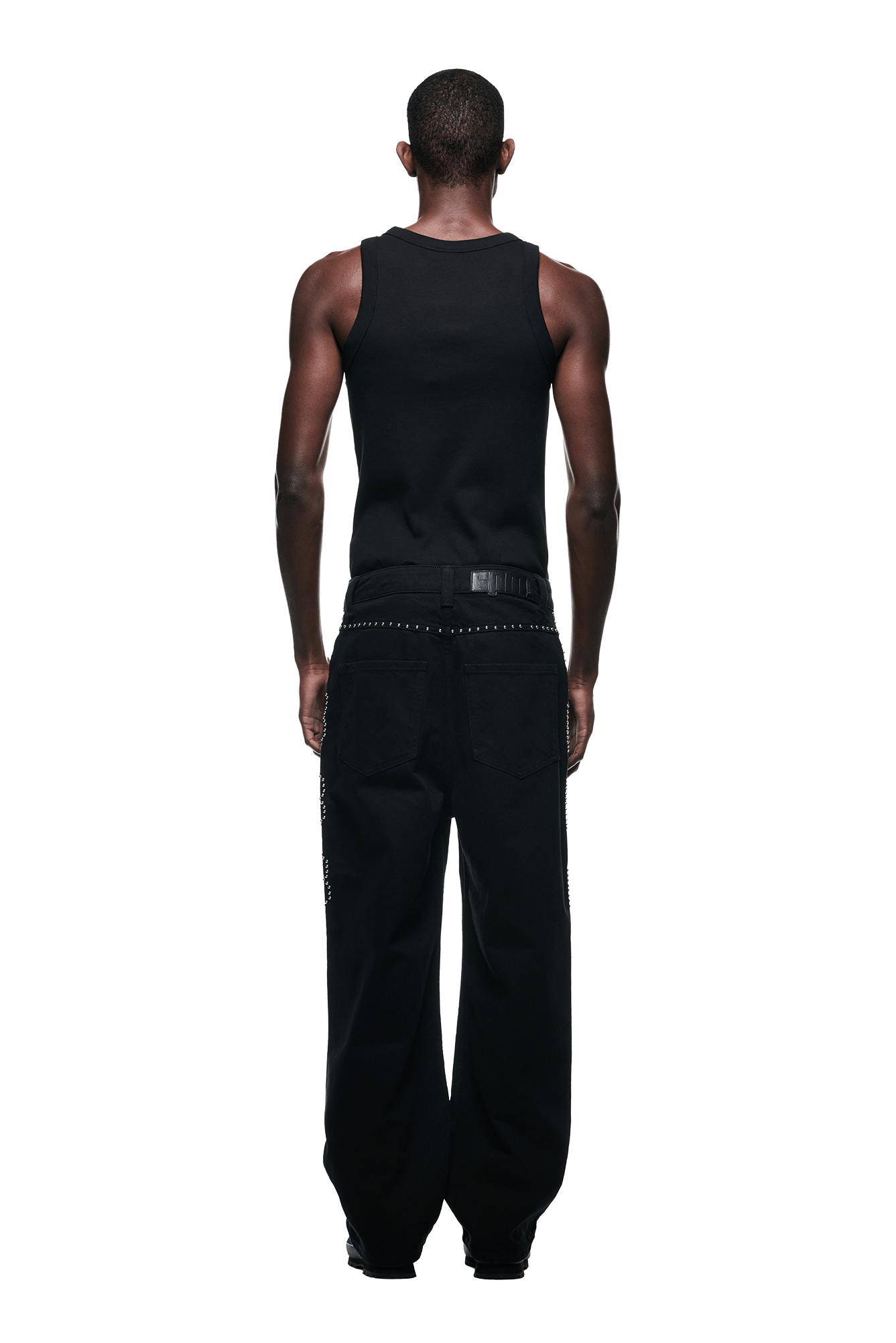 A man stands facing away from the camera in 6PM STUDS DENIM BLACK pants, paired with a black sleeveless tank top and black shoes, against a plain white background.