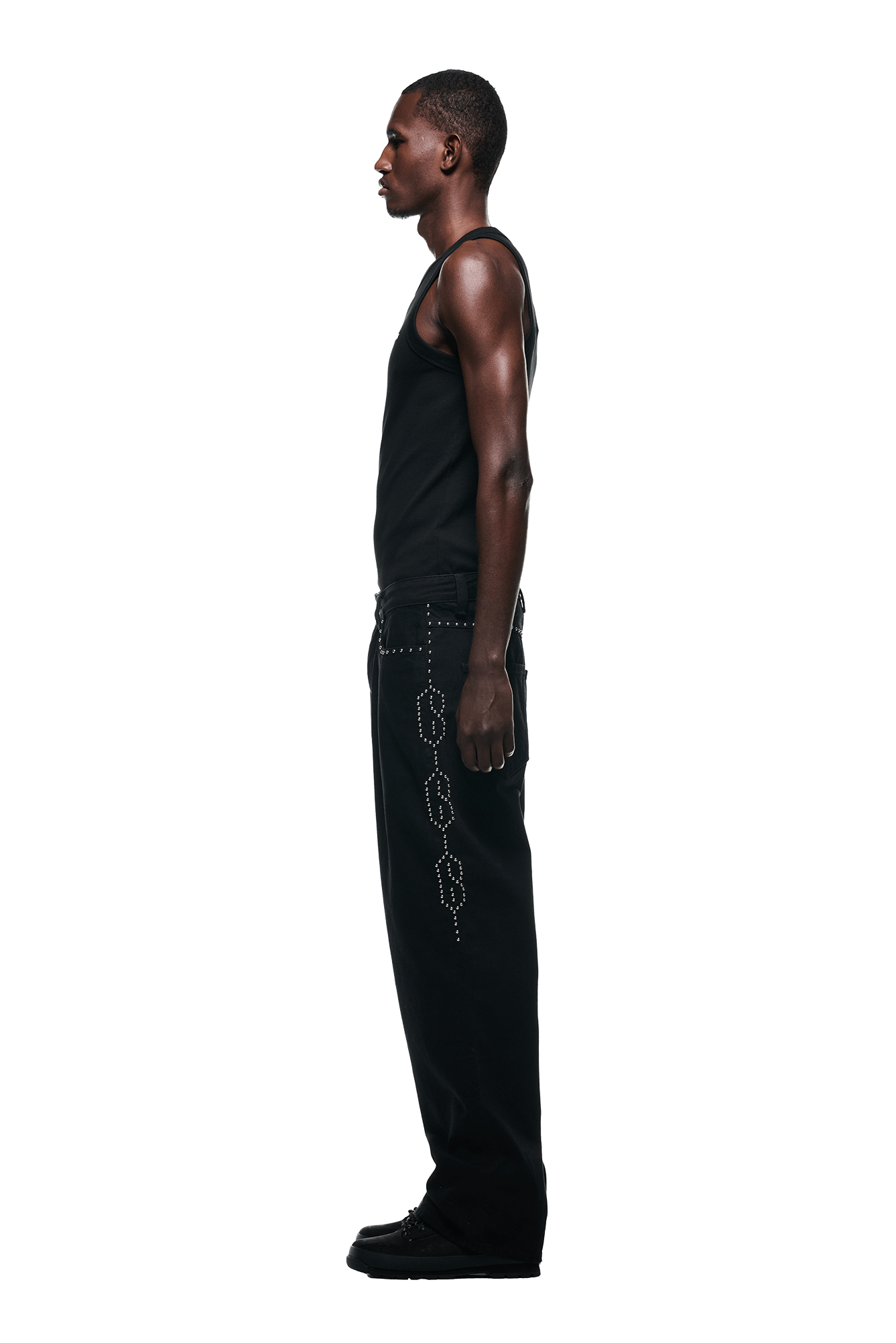 A man in profile faces left, wearing 6PM’s STUDS DENIM BLACK pants with a silver side pattern, a black tank top, and black shoes against a plain white background.