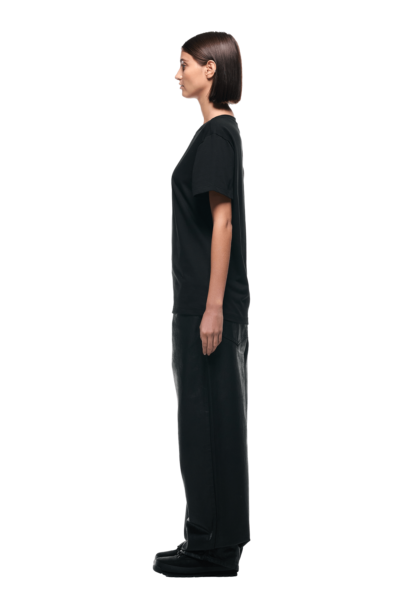 A woman with straight dark hair stands in profile, facing left, wearing the 6PM MONEY GUY TEE BLACK, loose black pants, and black shoes against a plain white background.