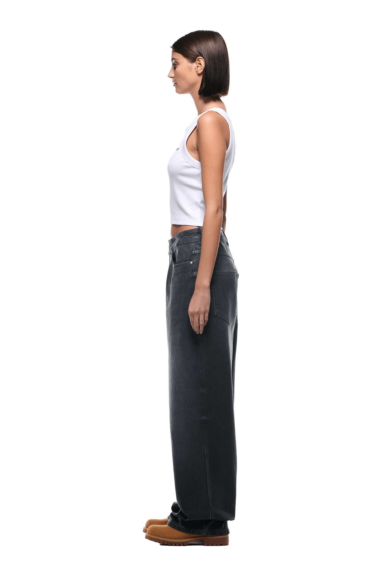 A woman with straight brown hair stands in profile, wearing a white tank top, 6PM WAX COATED DENIM GREY jeans, and brown shoes against a plain white background.