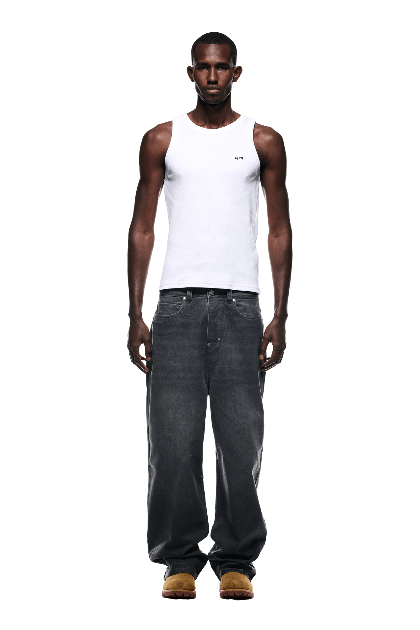 A man stands facing forward against a white background, wearing 6PM’s WAX COATED DENIM GREY jeans with a fitted white tank top and brown shoes.