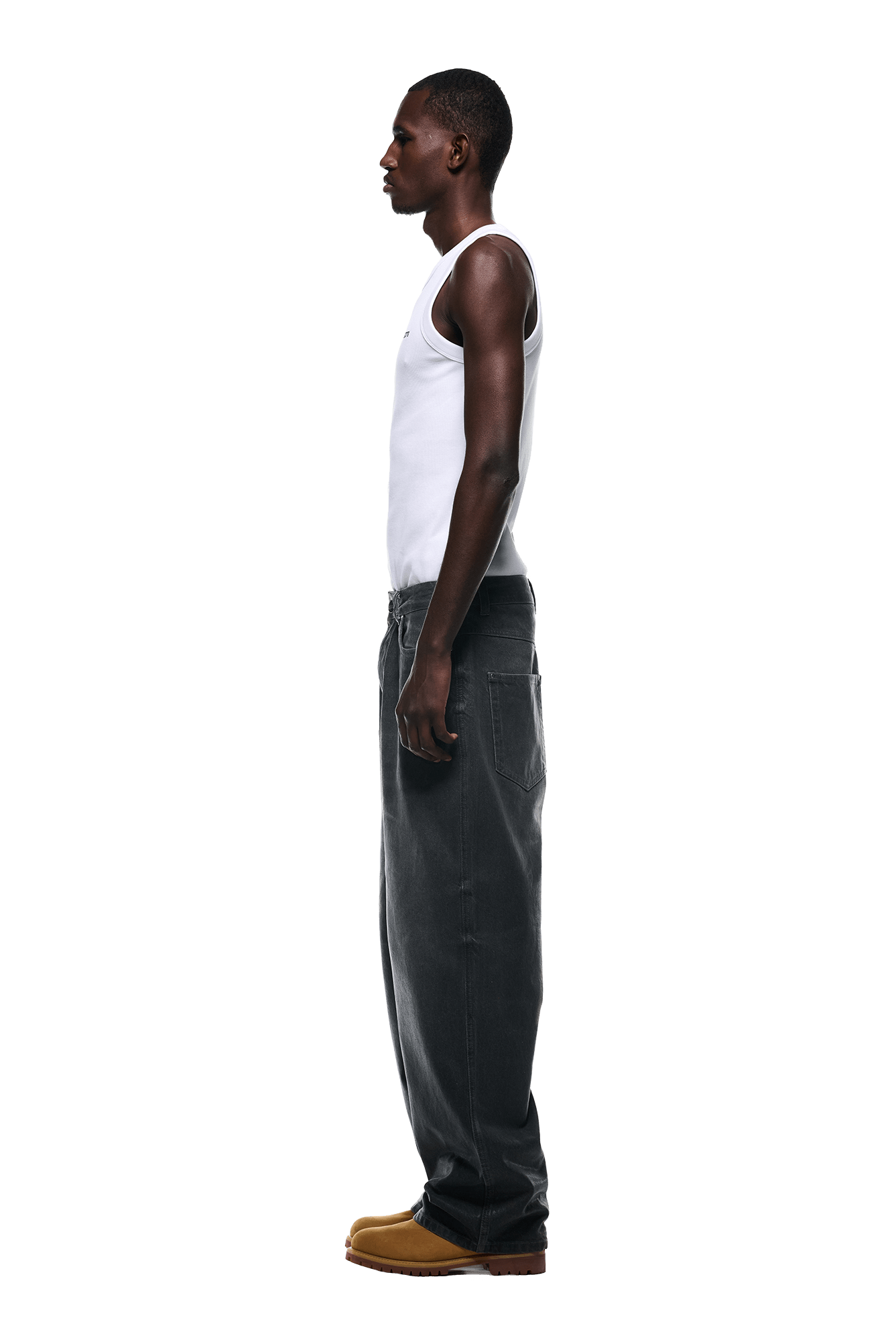A man stands in profile facing left, wearing a white sleeveless tank top, 6PM’s WAX COATED DENIM GREY jeans, and tan shoes against a plain white background.