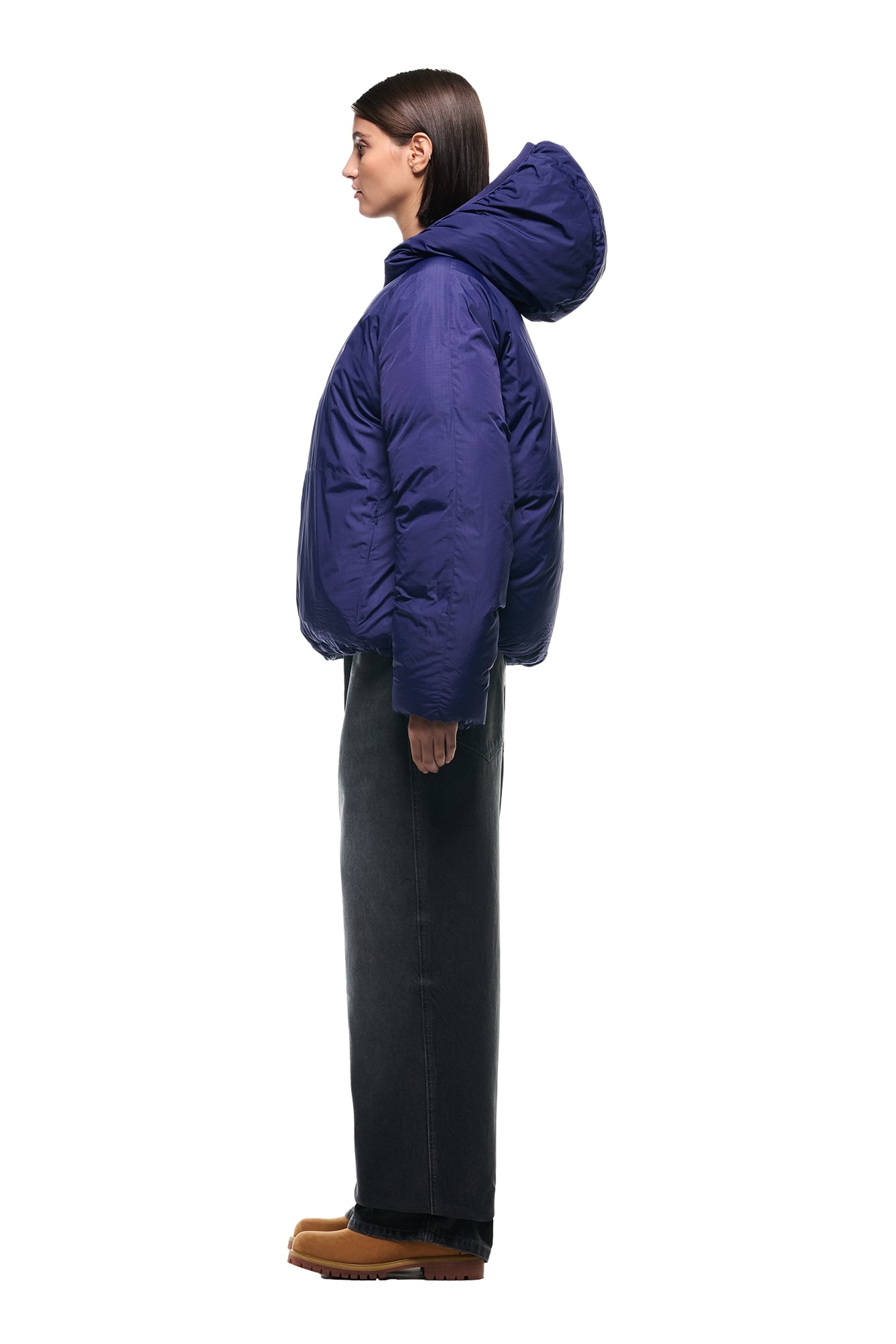 A person stands in profile against a white background, wearing the 6PM REVERSIBLE PUFFER JACKET BLUE with a hood, paired with dark straight-leg pants and tan shoes.