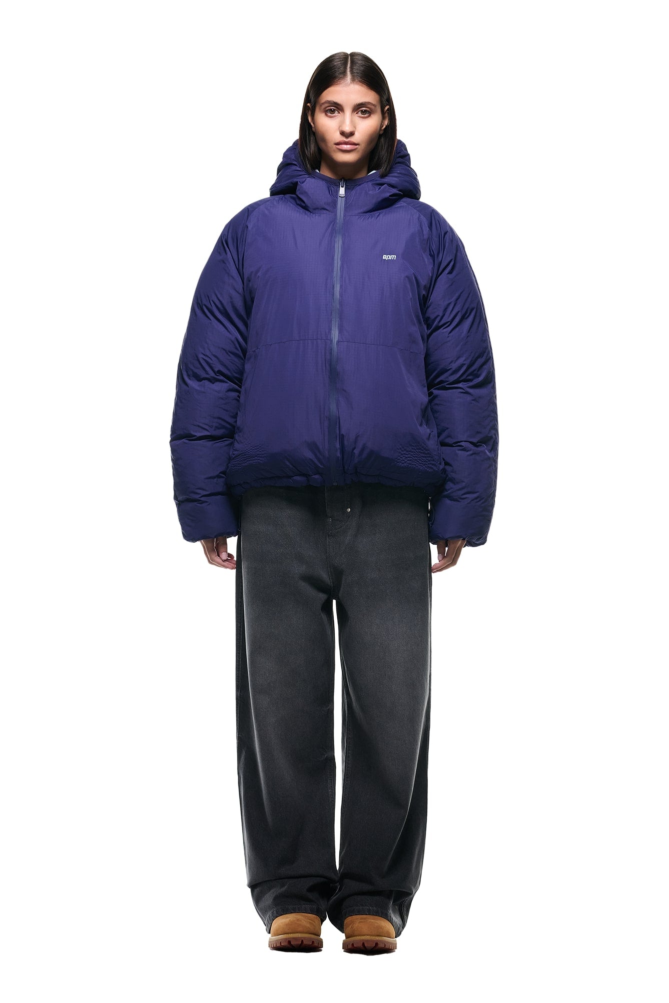 A person stands facing forward, wearing the 6PM REVERSIBLE PUFFER JACKET BLUE, wide-leg dark gray jeans, and brown shoes against a plain white background.