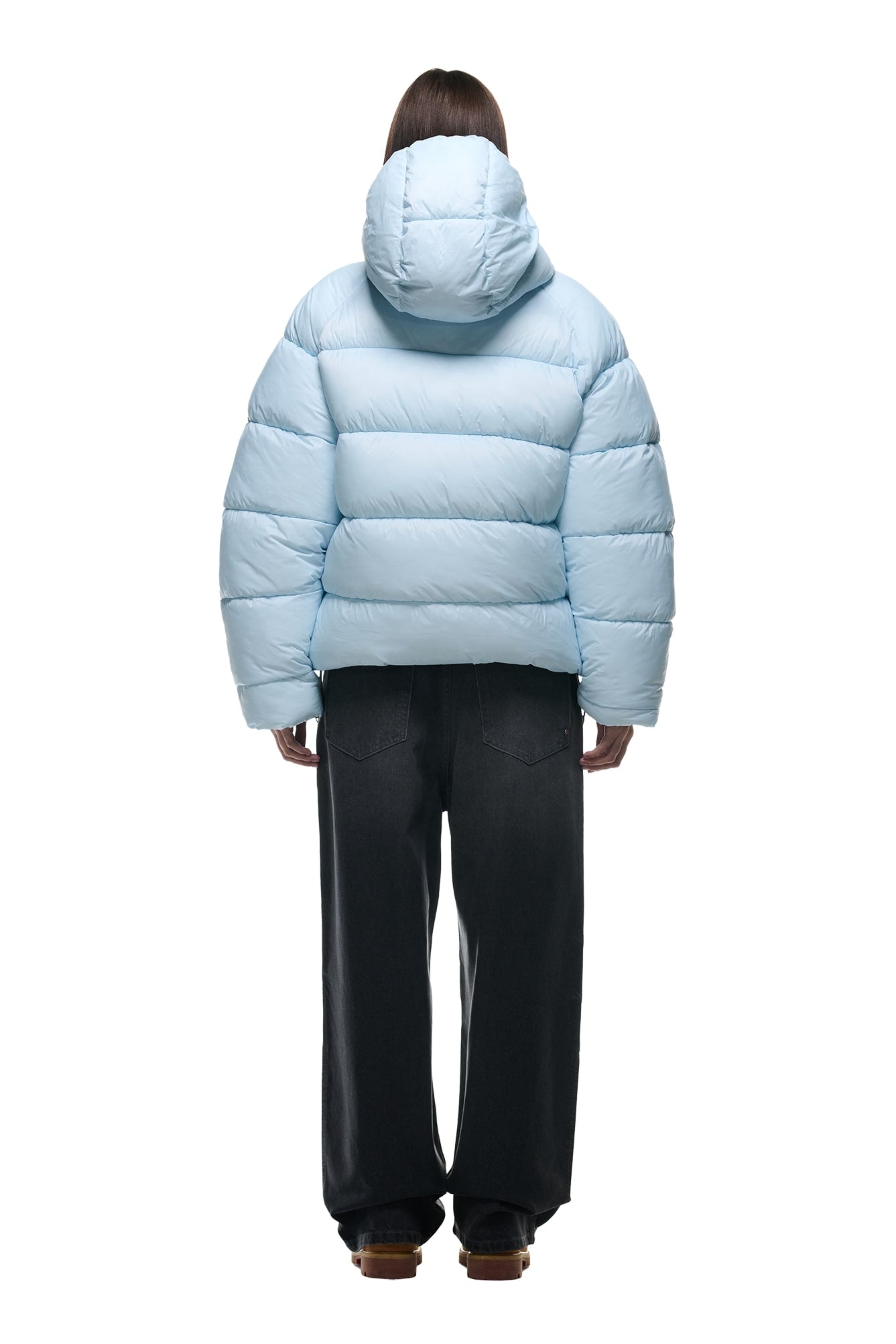 A person stands facing away, wearing the 6PM REVERSIBLE PUFFER JACKET BLUE with a hood and loose-fitting dark jeans against a white background.