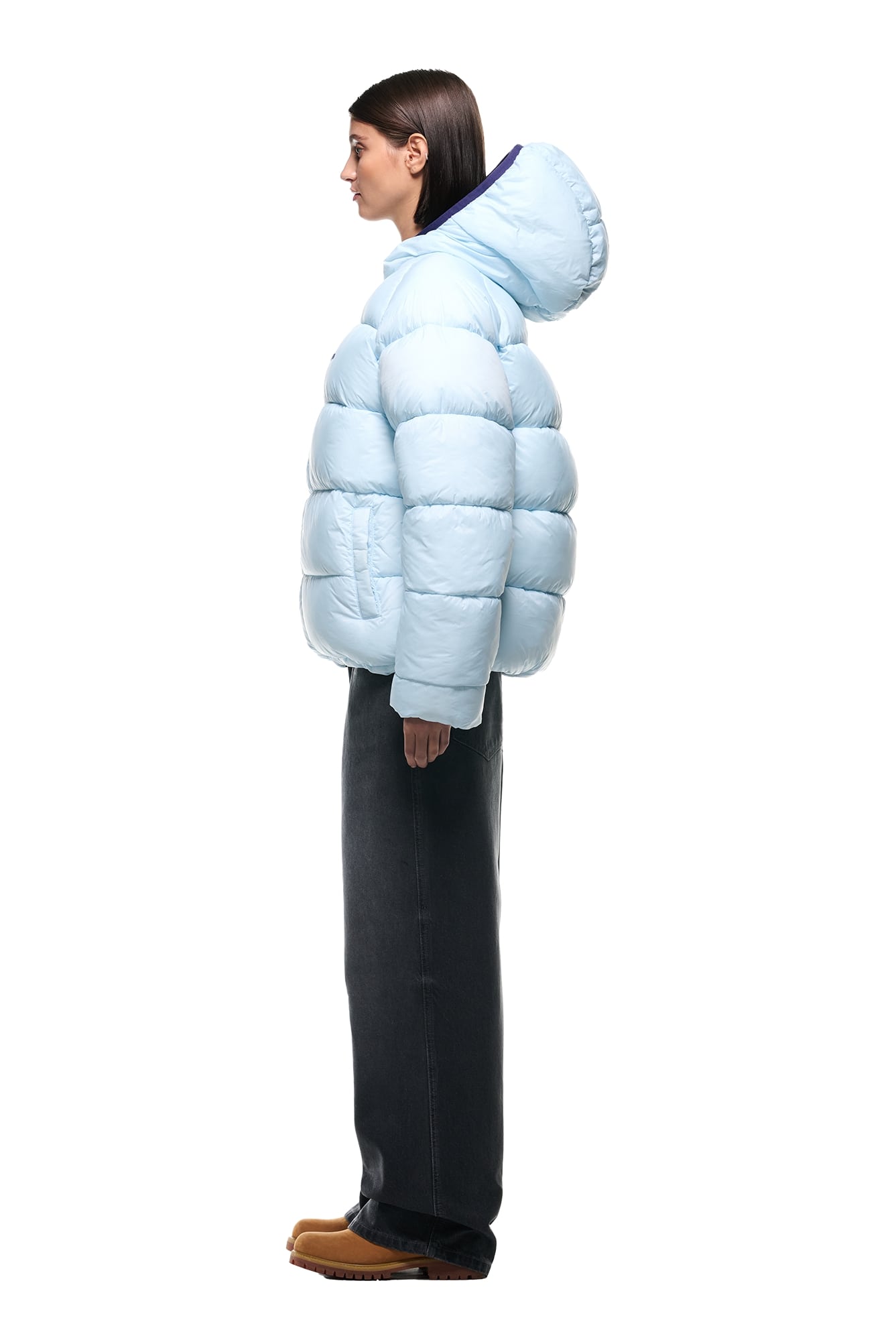 A woman stands in profile against a white background, wearing the 6PM REVERSIBLE PUFFER JACKET BLUE with a hood, dark straight-leg pants, and brown shoes.