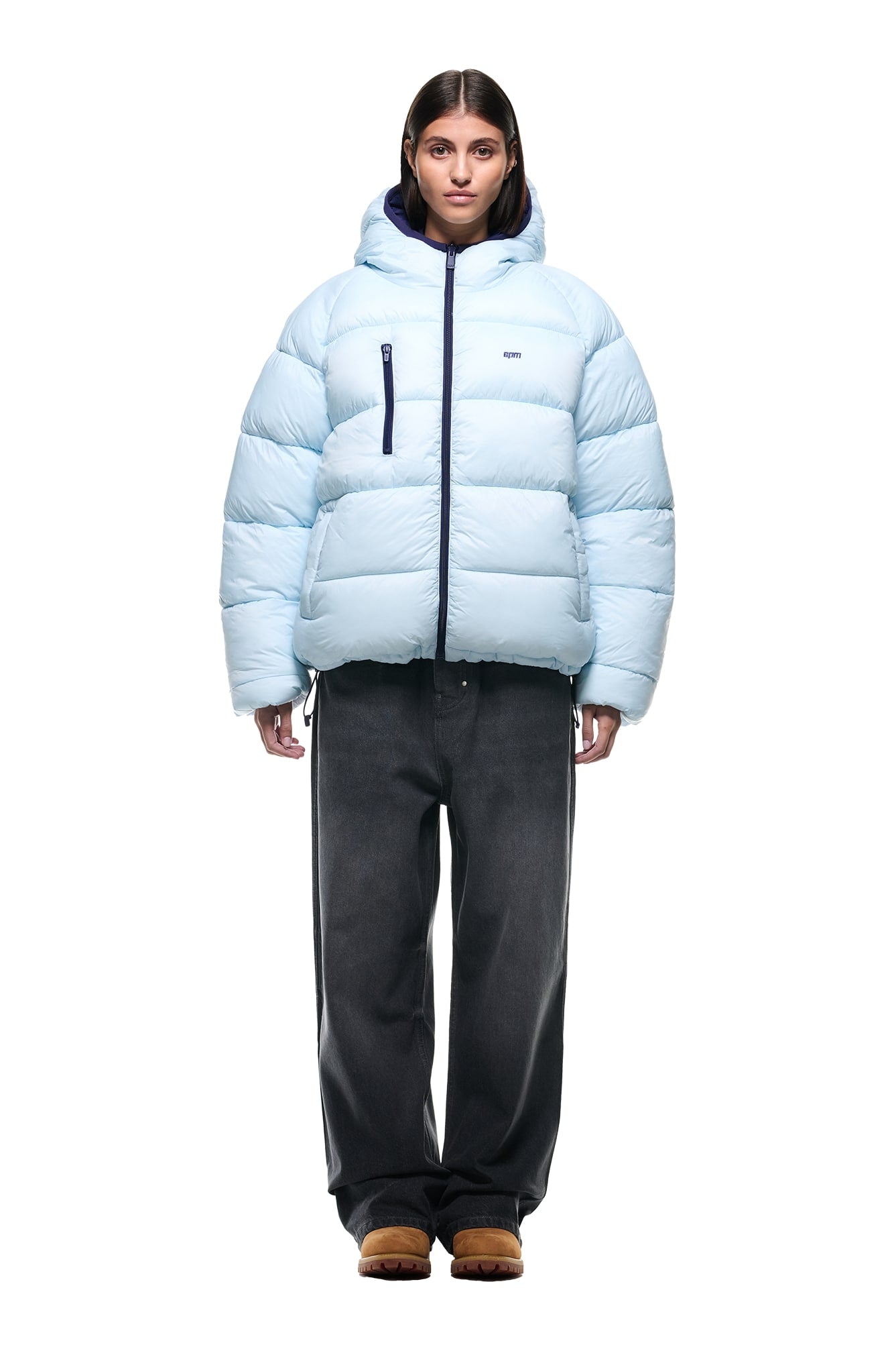 A person stands facing forward against a plain white background, wearing the 6PM REVERSIBLE PUFFER JACKET BLUE with a hood, paired with loose dark gray pants and brown shoes.