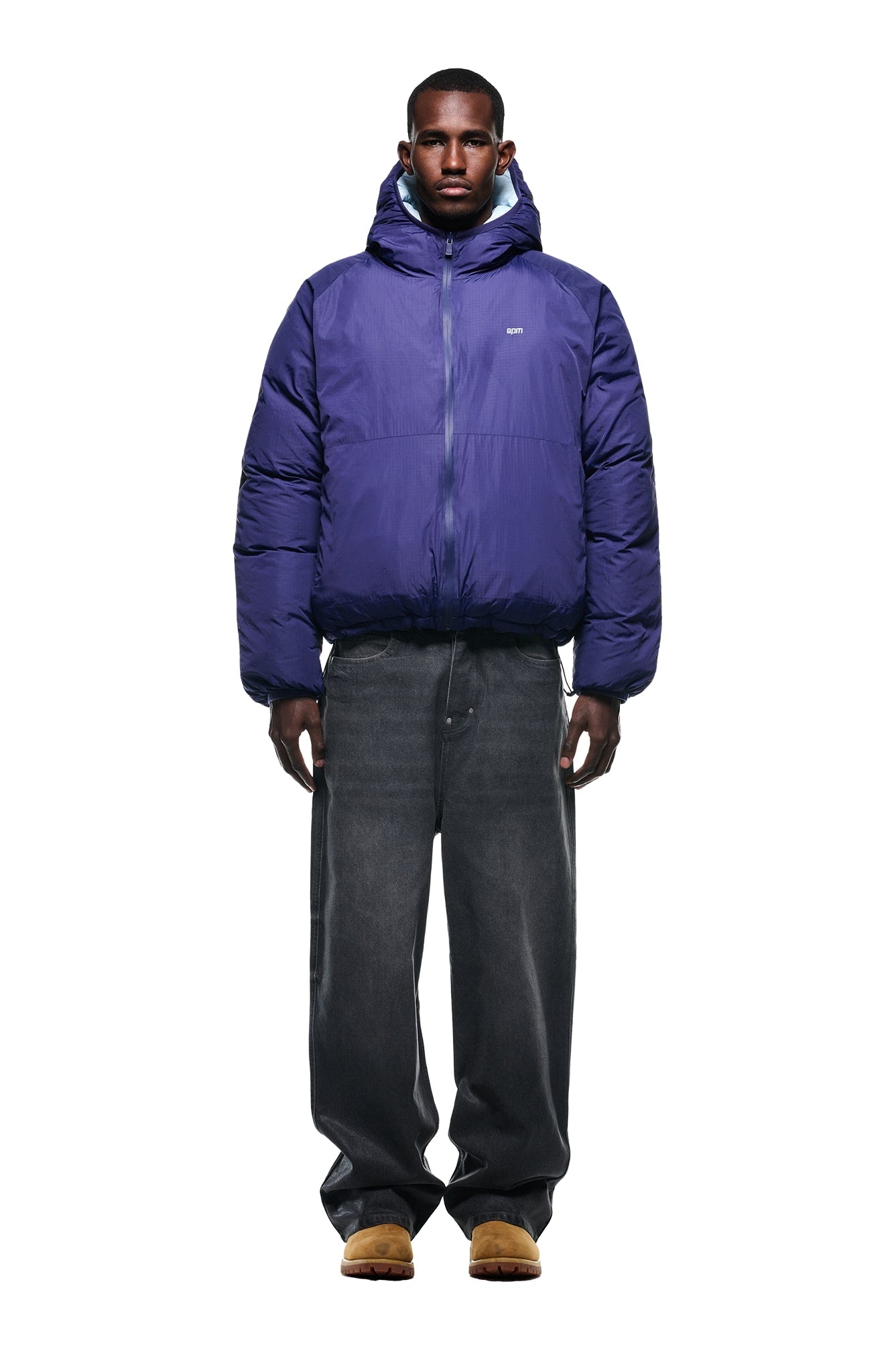 A man stands facing forward wearing the 6PM REVERSIBLE PUFFER JACKET BLUE with a hood, loose-fitting dark gray jeans, and tan suede shoes against a plain white background.