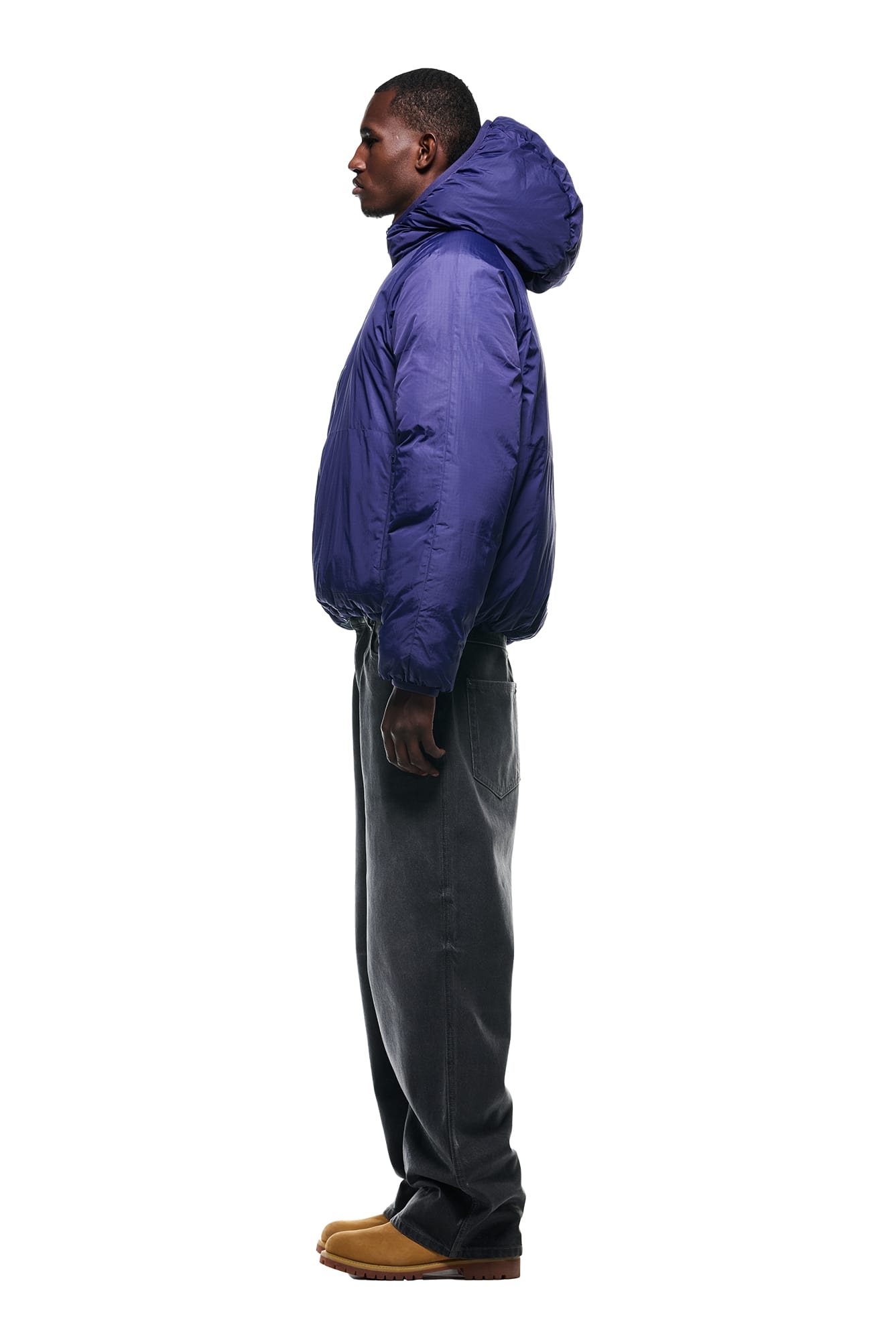 A person stands in profile against a white background, wearing the 6PM REVERSIBLE PUFFER JACKET BLUE, loose dark gray jeans, and tan shoes.
