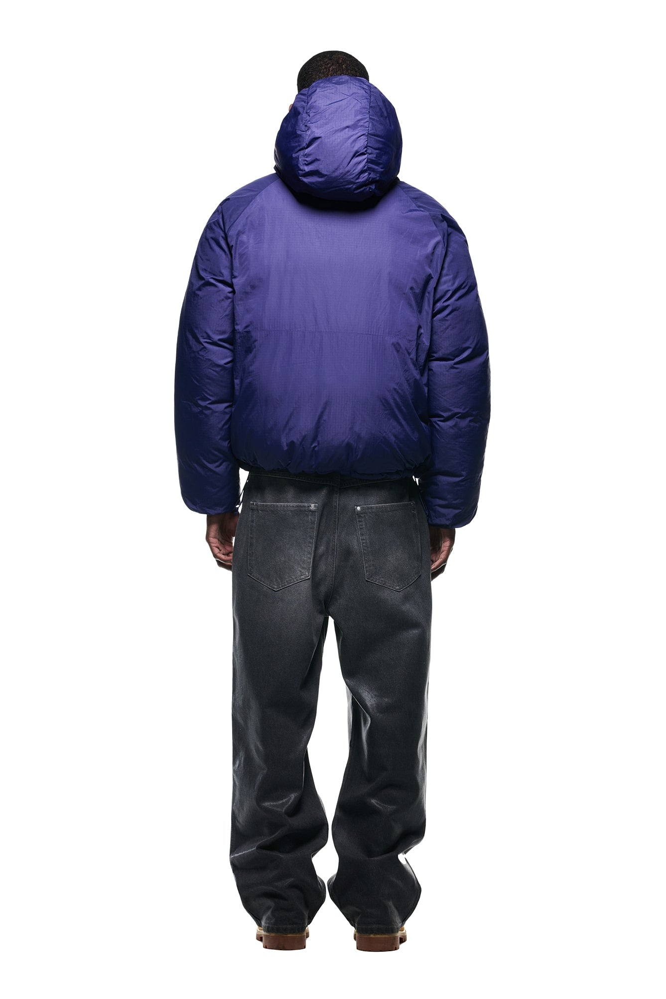 A person stands facing away, wearing the 6PM REVERSIBLE PUFFER JACKET BLUE with a hood, paired with loose dark gray jeans and brown shoes, against a plain white background.