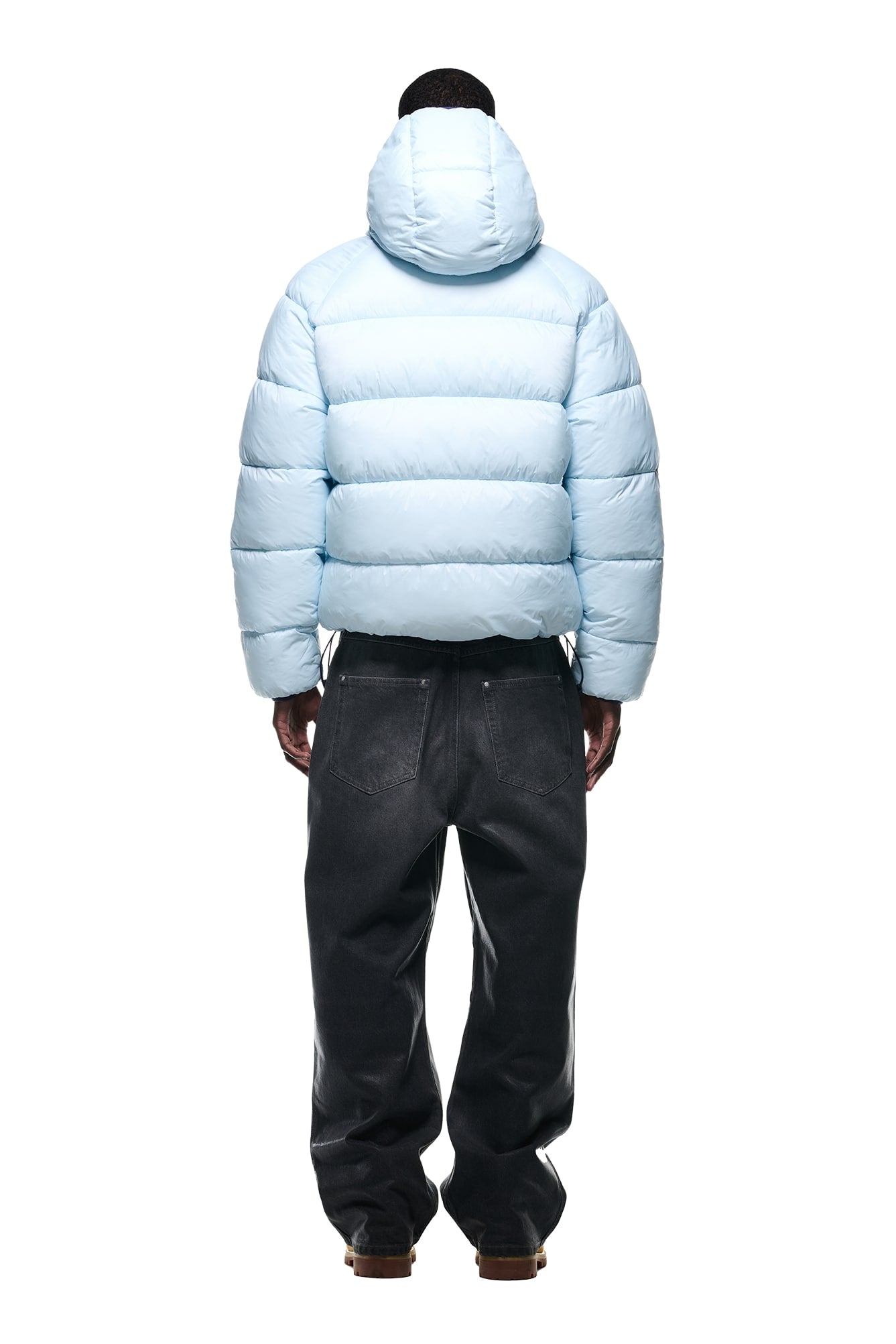 A person stands facing away, wearing the 6PM REVERSIBLE PUFFER JACKET BLUE with a hood and loose-fitting dark jeans against a white background.