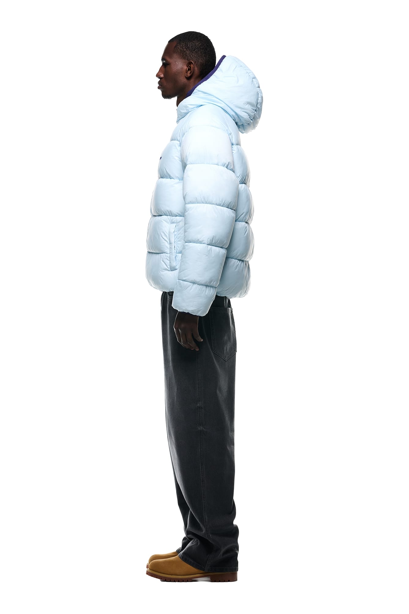 A man in profile faces left, wearing the 6PM REVERSIBLE PUFFER JACKET BLUE with a hood, dark pants, and tan shoes, against a plain white background.