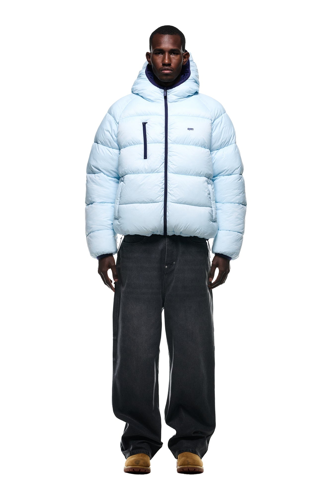 A person stands facing forward wearing the 6PM REVERSIBLE PUFFER JACKET BLUE, paired with loose dark gray jeans and brown shoes, against a white background.