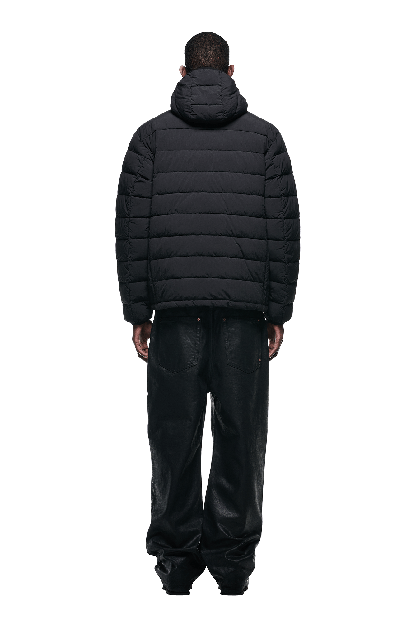 A person stands with their back to the camera, dressed in the 6PM LIGHT PUFFER JACKET BLACK with a hood and loose-fitting black pants against a plain white background.