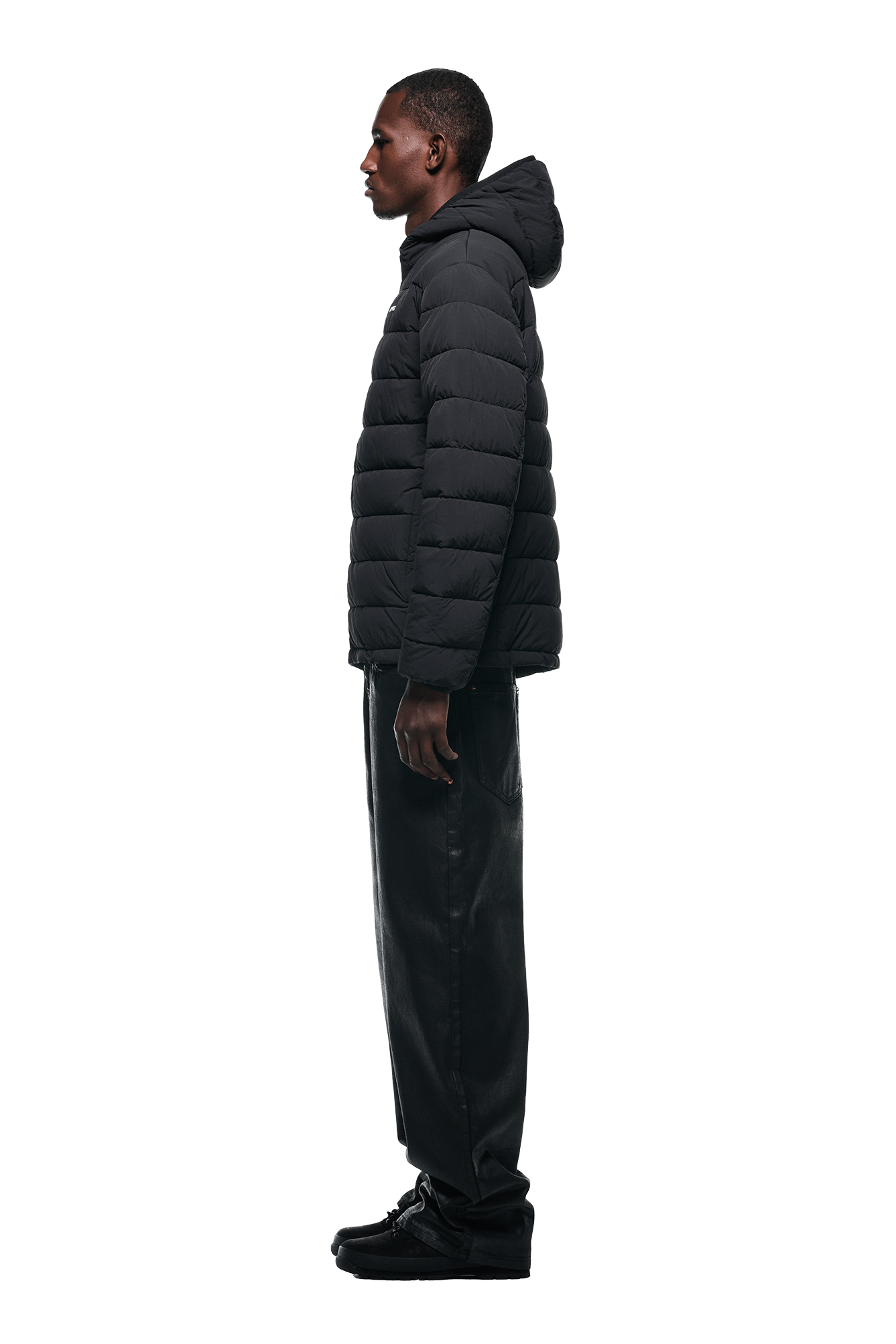 A man in profile wears the 6PM LIGHT PUFFER JACKET BLACK with black pants and shoes, standing against a plain white background.