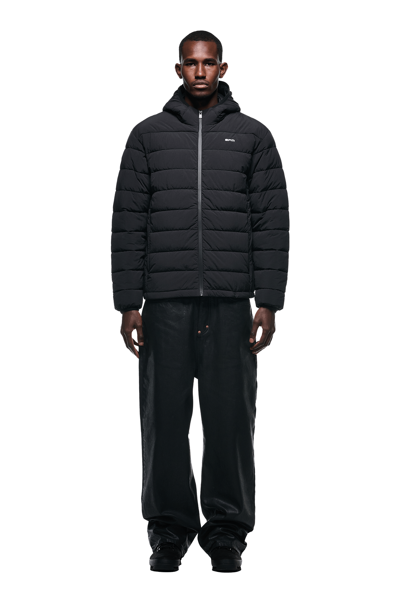 A man faces forward against a plain white background, wearing the 6PM LIGHT PUFFER JACKET BLACK, loose black pants, and black sneakers.