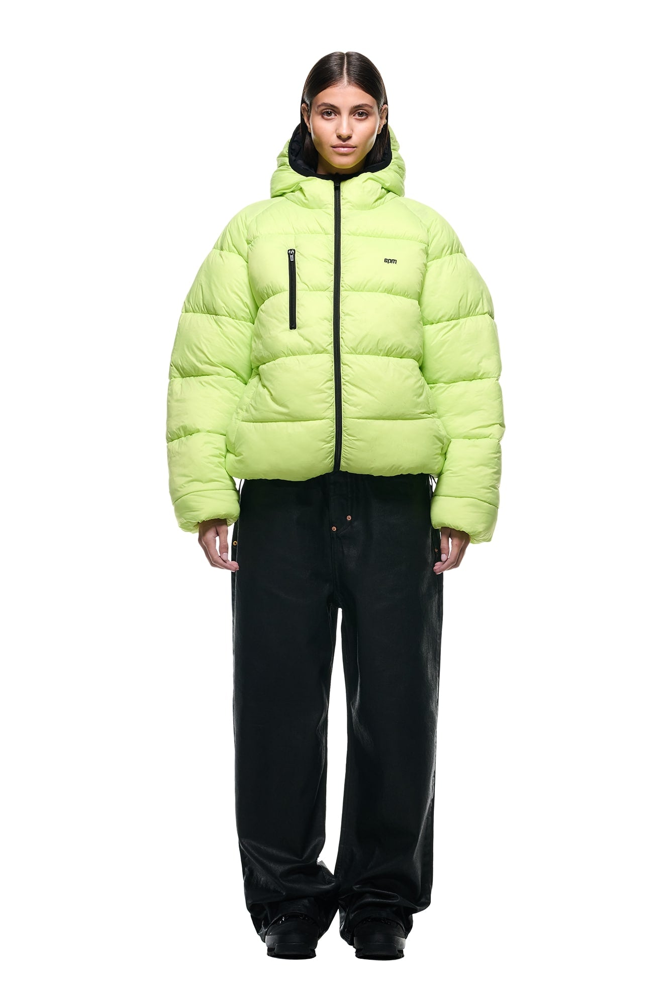 A person stands facing forward in the 6PM REVERSIBLE PUFFER JACKET BLACK with a hood and large front zipper pocket, paired with loose black pants and black shoes against a plain white background.