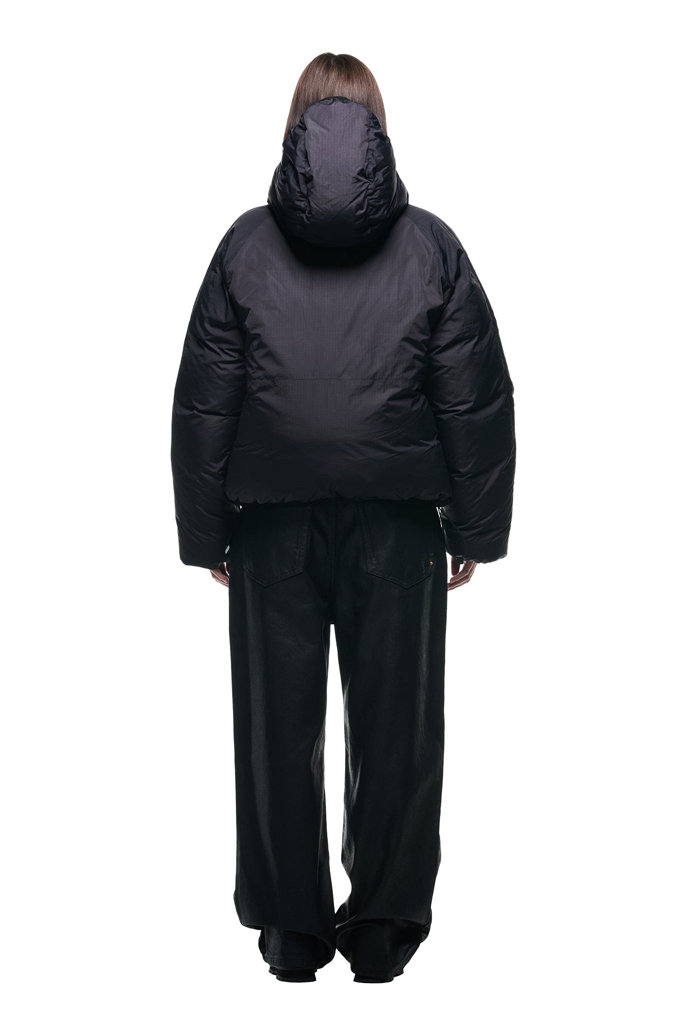 A person stands with their back to the camera, wearing the 6PM REVERSIBLE PUFFER JACKET BLACK with a hood and loose-fitting black pants against a white background.