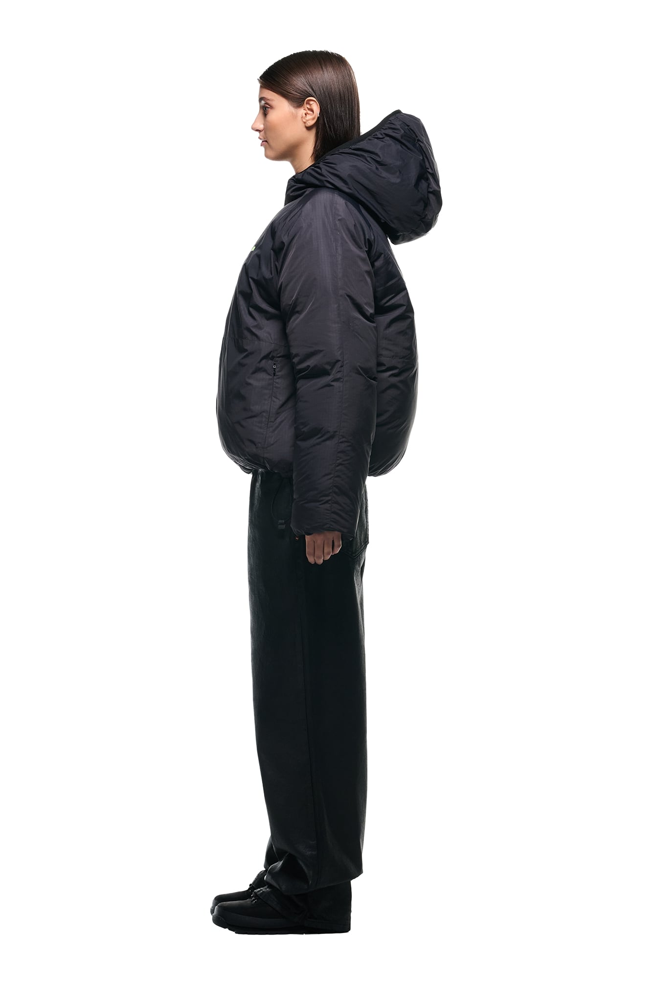 A woman stands in profile against a white background, wearing the 6PM REVERSIBLE PUFFER JACKET BLACK with a hood, paired with black wide-leg pants and black shoes.