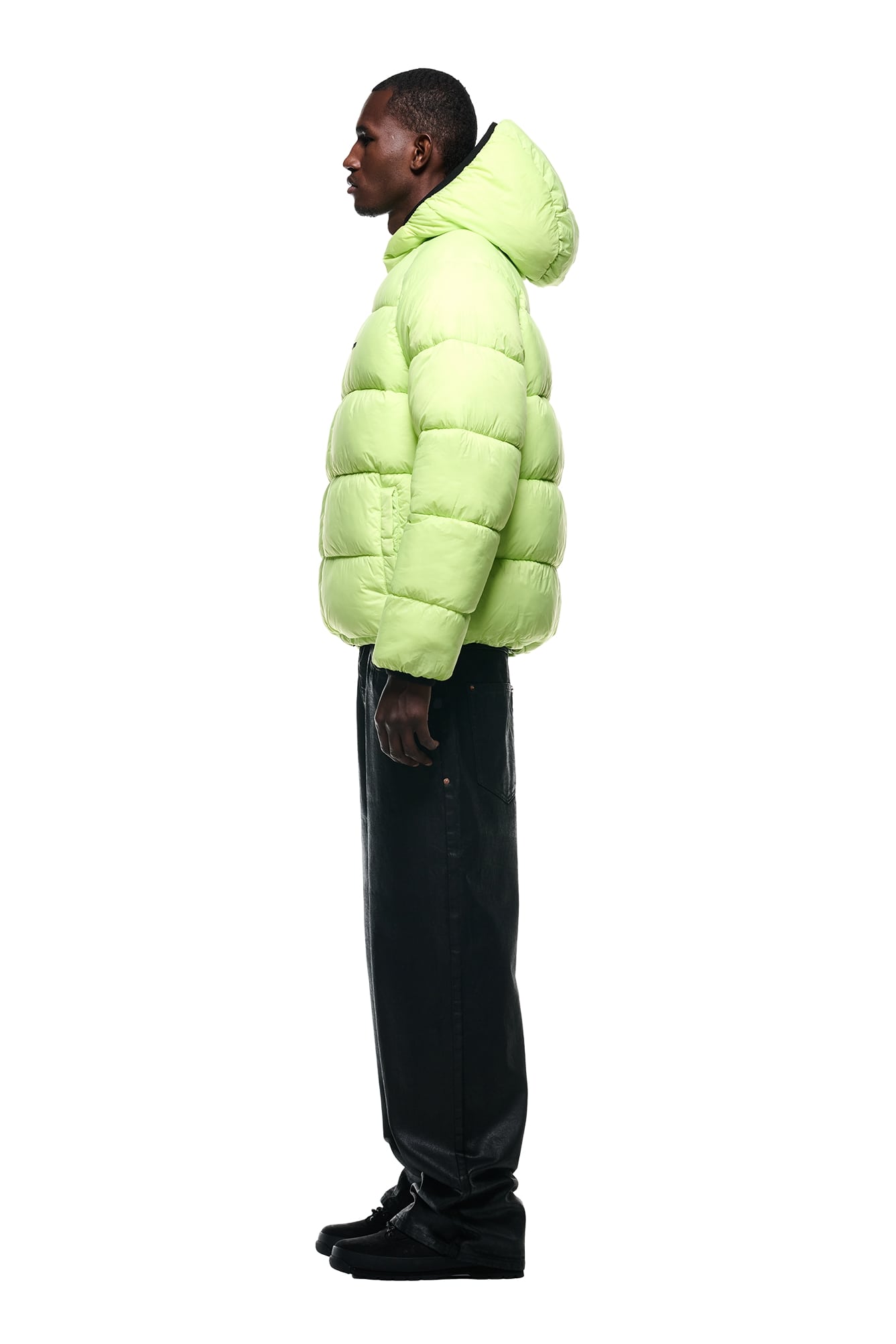 A person stands in profile against a white background, wearing the 6PM REVERSIBLE PUFFER JACKET BLACK, along with loose black pants and black shoes.