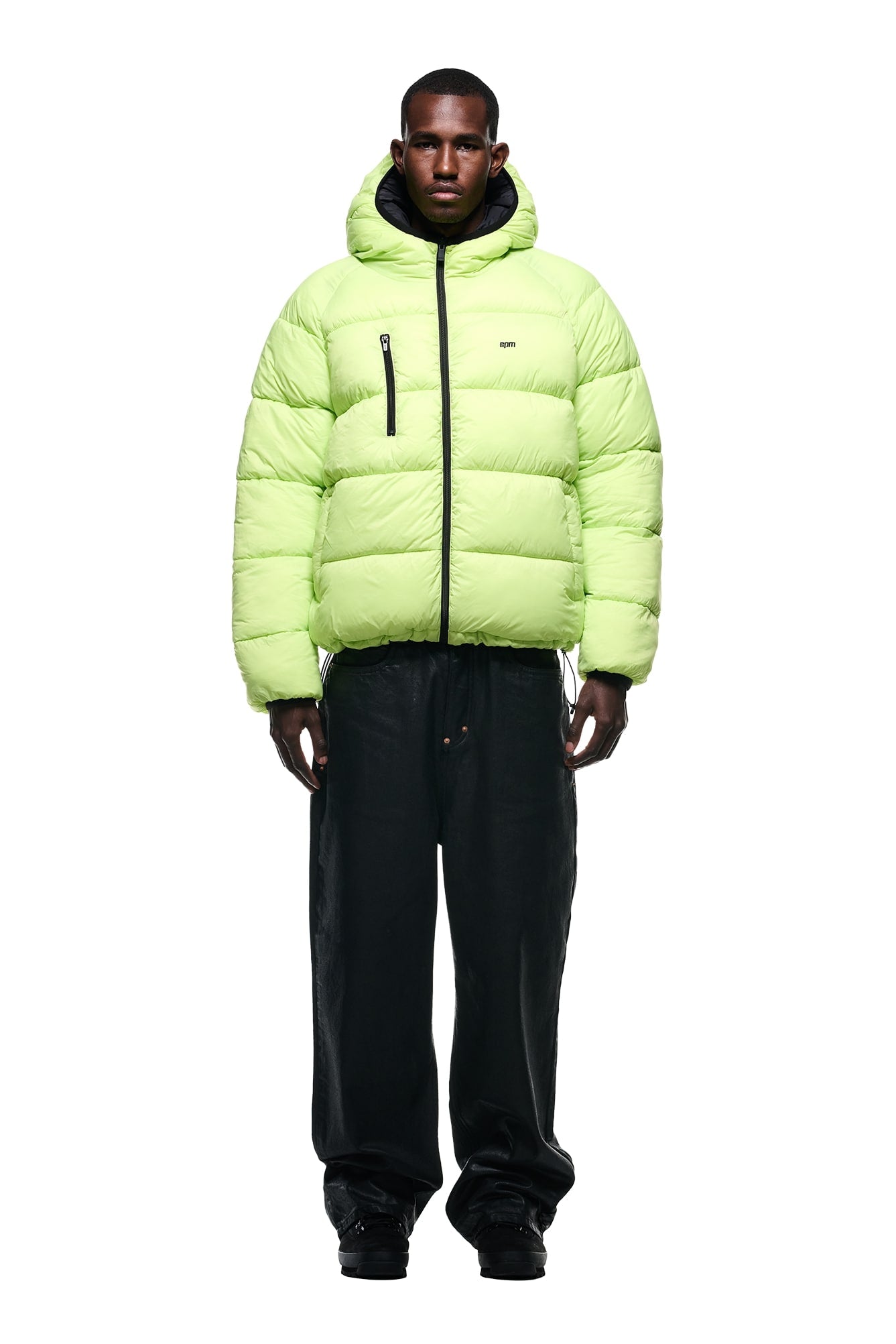 A person faces forward, wearing the 6PM REVERSIBLE PUFFER JACKET BLACK with a hood and loose black pants against a plain white background.
