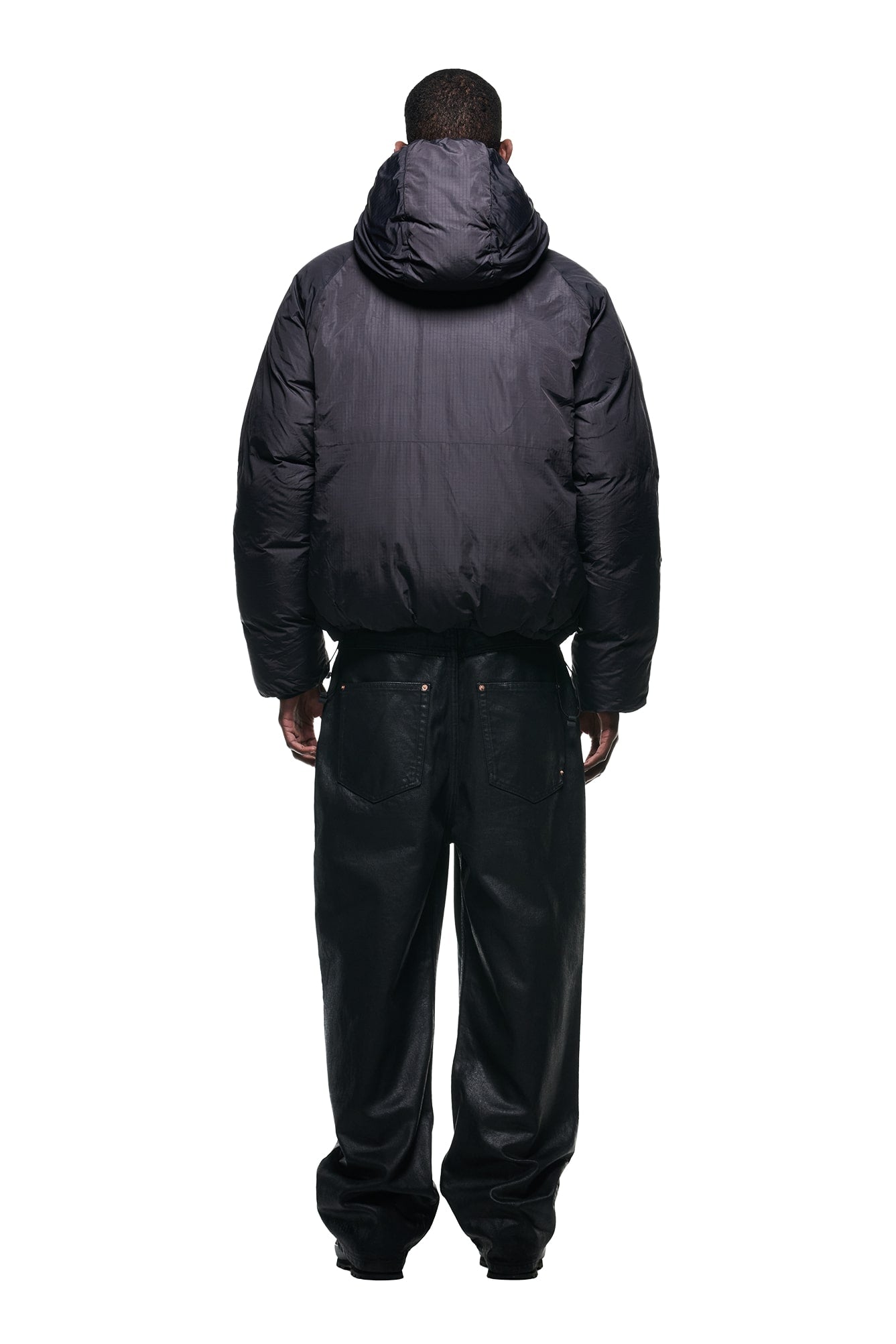 A person faces away from the camera in a 6PM REVERSIBLE PUFFER JACKET BLACK with a hood and loose black pants, set against a plain white background.