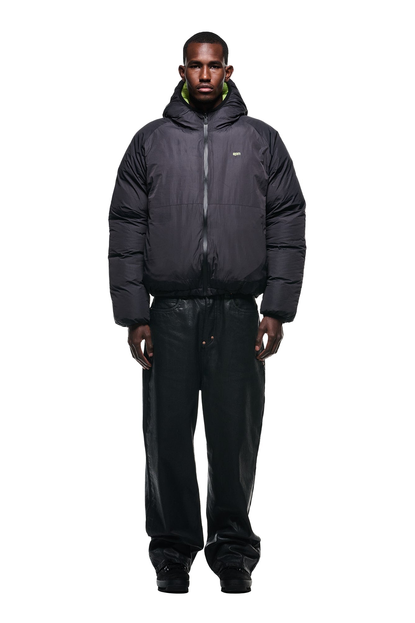 A man faces forward against a plain white background, wearing the 6PM REVERSIBLE PUFFER JACKET BLACK with a hood, paired with black wide-leg pants and black shoes.